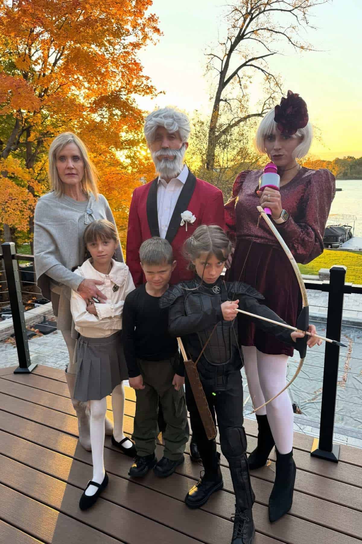 A group of adults and children in costumes pose outdoors on a deck in front of autumn trees and a lake at sunset.