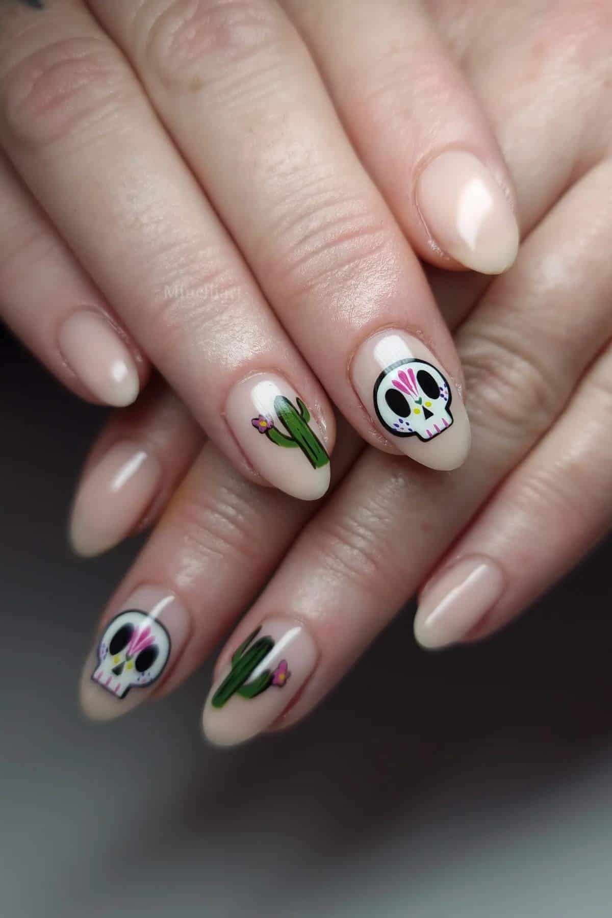 Hands with almond-shaped nude nails featuring sugar skull and cactus nail art on the ring and middle fingers.