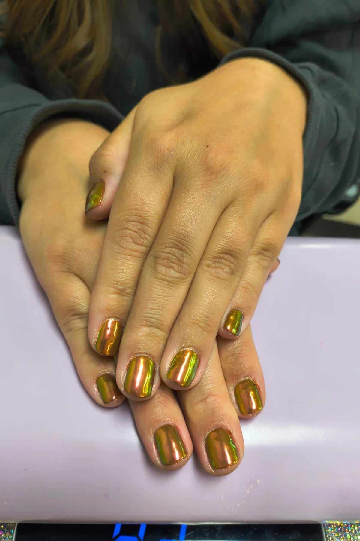 Two hands rest on a light purple surface, displaying short fingernails painted with a shiny, metallic gold-green polish.