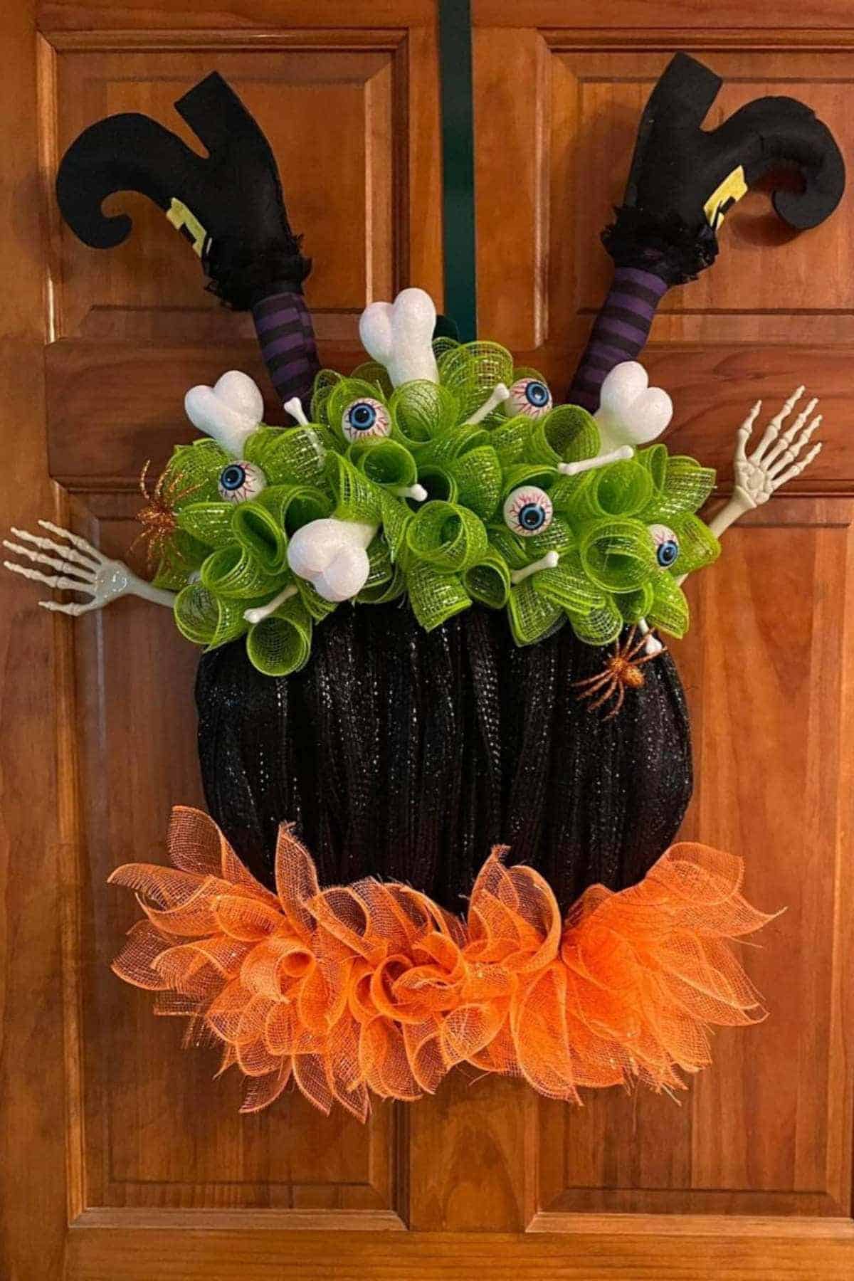 Halloween wreath with witch legs, green mesh flowers, eyeball decorations, skeleton hands, white bones, orange mesh, and small spiders, hanging on a wooden door.
