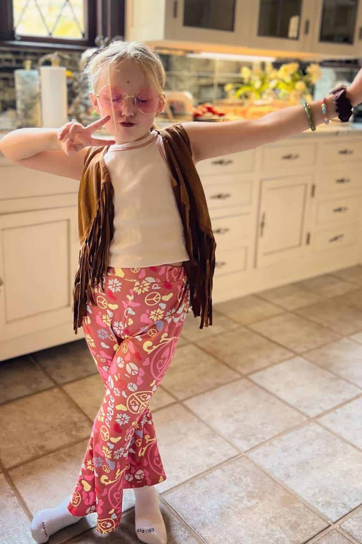 Child in a kitchen wearing peace sign sunglasses, a fringed vest, and pink floral bell-bottom pants, posing with a peace sign hand gesture.