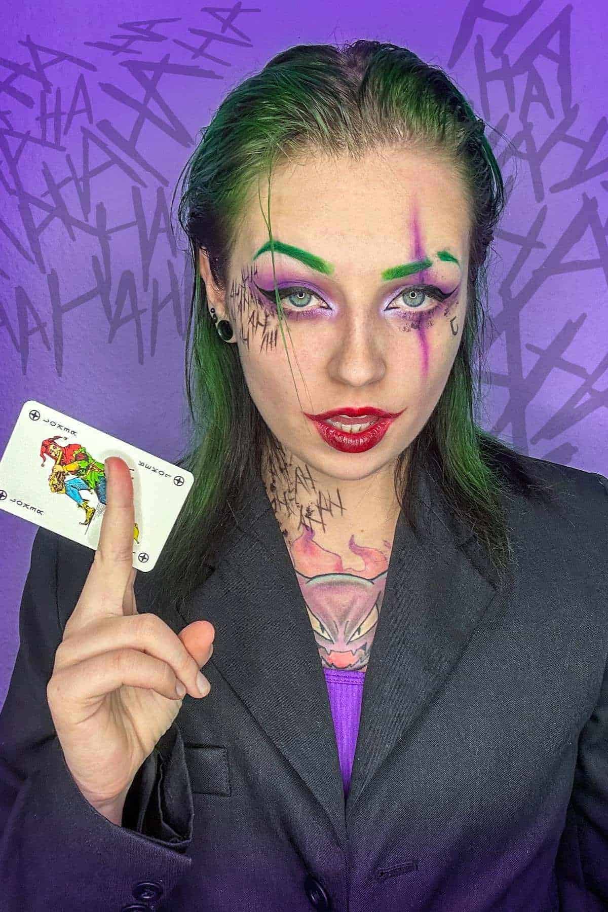 Person with green hair and Joker-inspired makeup holds up a Joker card, wearing a black suit, with tally marks and purple “HA” graffiti on a purple background.