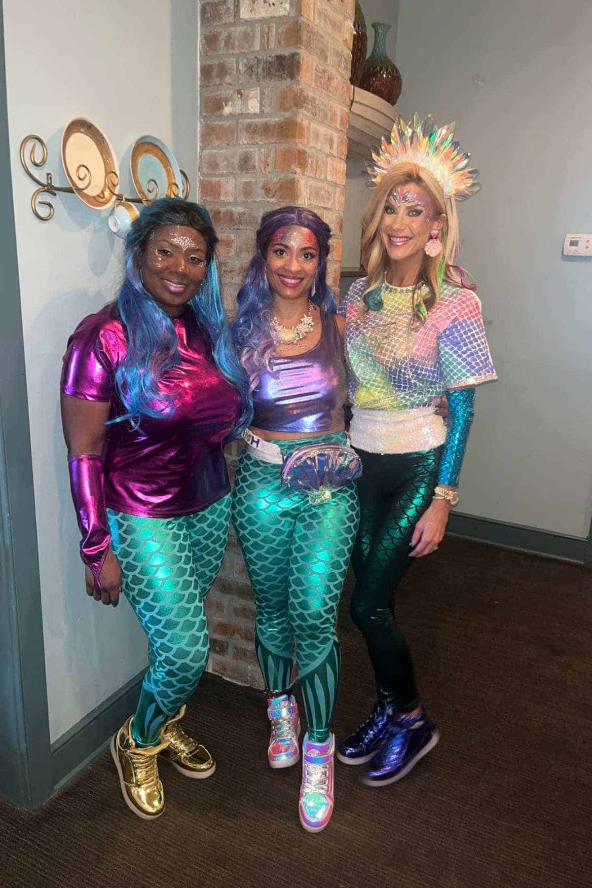 Three people stand indoors wearing colorful mermaid-themed costumes with shiny tops, metallic leggings, and vibrant wigs, posing and smiling for the camera.