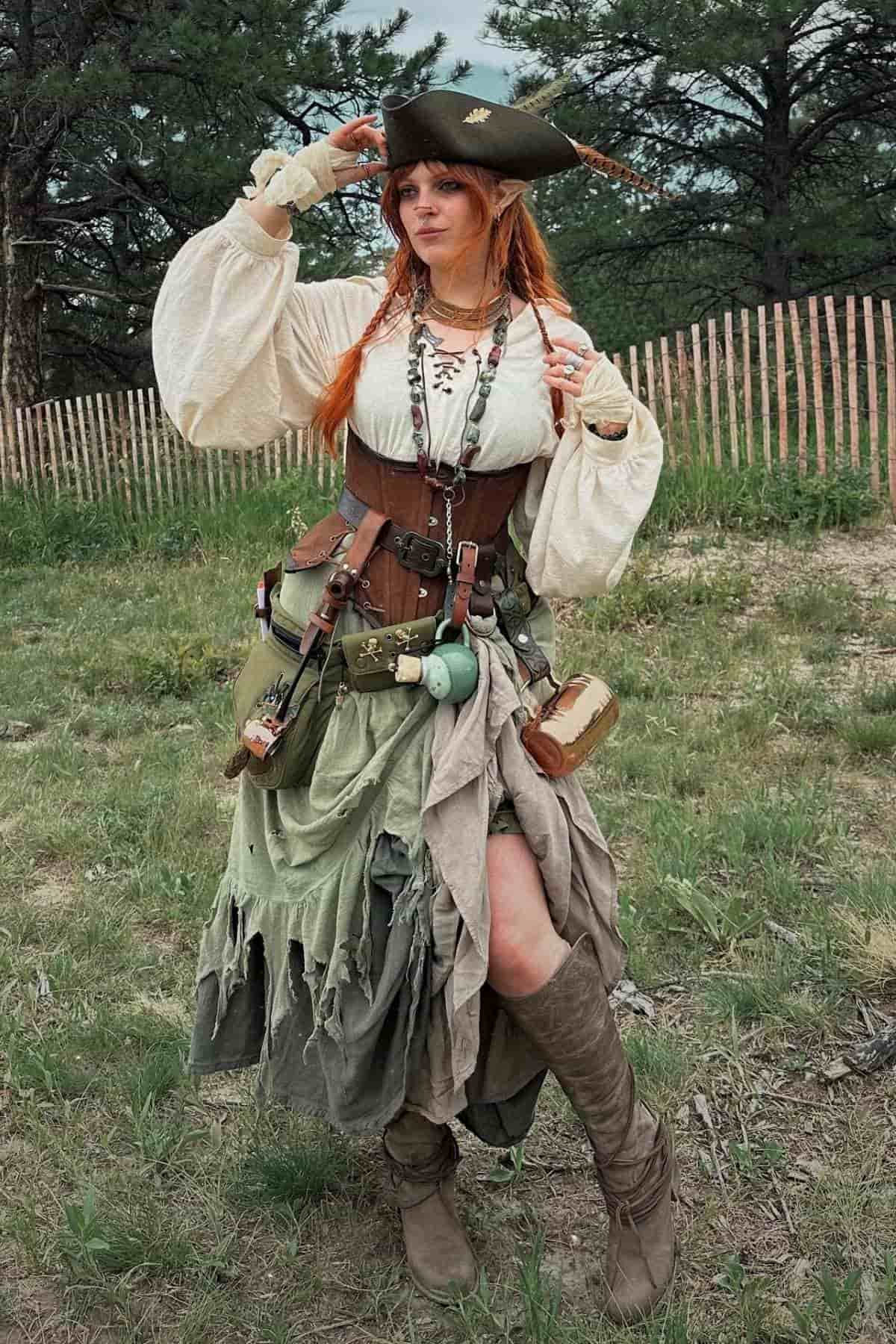 Person in a pirate costume stands outdoors on grass, wearing a tricorn hat, layered dress, boots, and accessories, holding one hand to their hat, with trees and a wooden fence in the background.