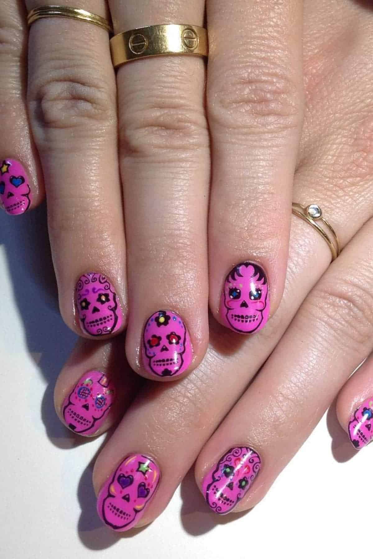 Hands with gold rings displaying pink nail art featuring decorative sugar skull designs with black outlines, hearts, and stars.