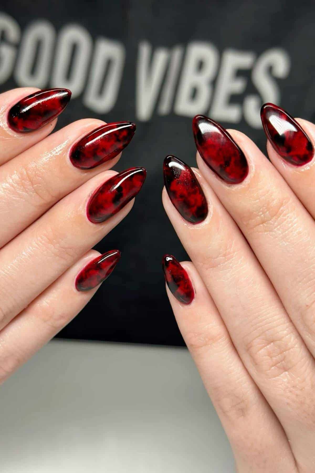 Hands with long, almond-shaped nails featuring a glossy red and black marbled design. The background has blurred text that reads "GOOD VIBES.