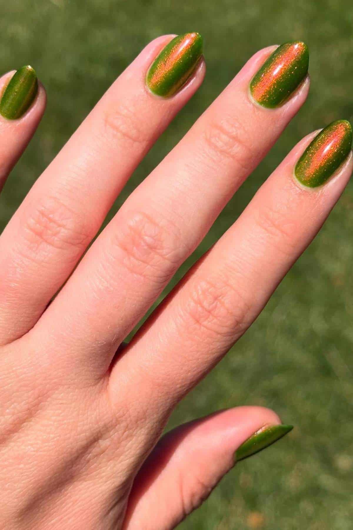 A hand with short, almond-shaped nails painted green with orange shimmer, shown against a blurred grassy background.