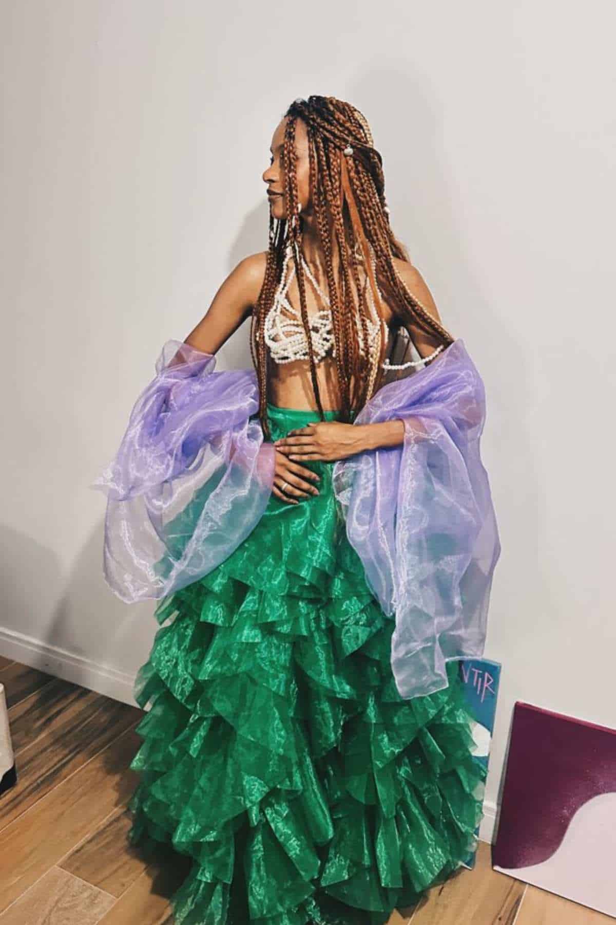 Person stands indoors wearing a green ruffled skirt, a crochet top, and a sheer lavender shawl. They have long braided hair and look to the side.