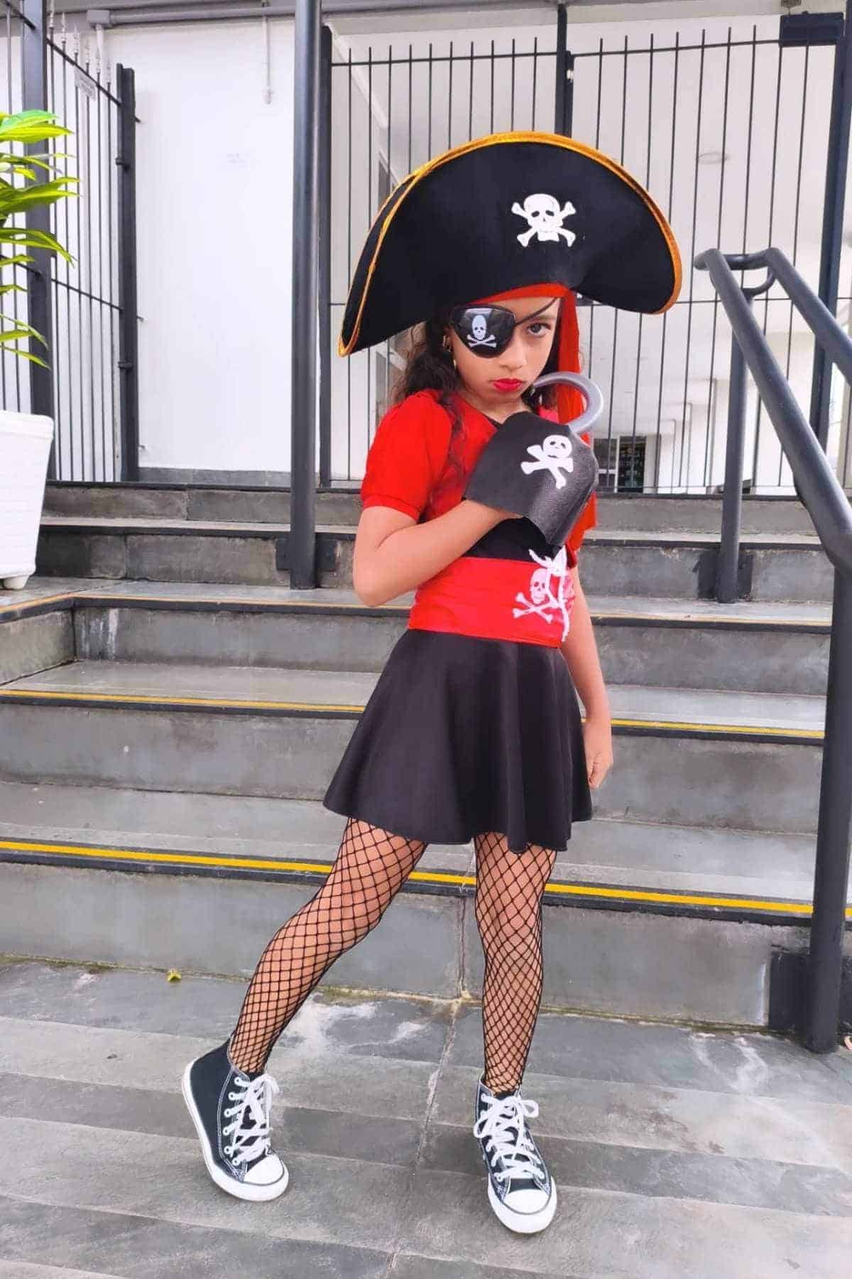 Child in a pirate costume with an eye patch, hat, and hook stands on steps, wearing a red shirt, black skirt, fishnet tights, and sneakers.