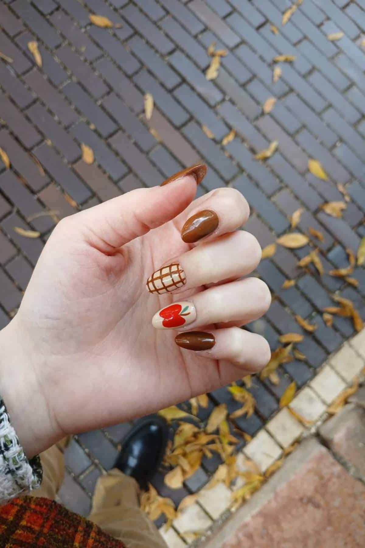 A hand with manicured nails painted in brown tones, one with a grid pattern and another with a red heart, is shown against a leaf-covered brick pavement.