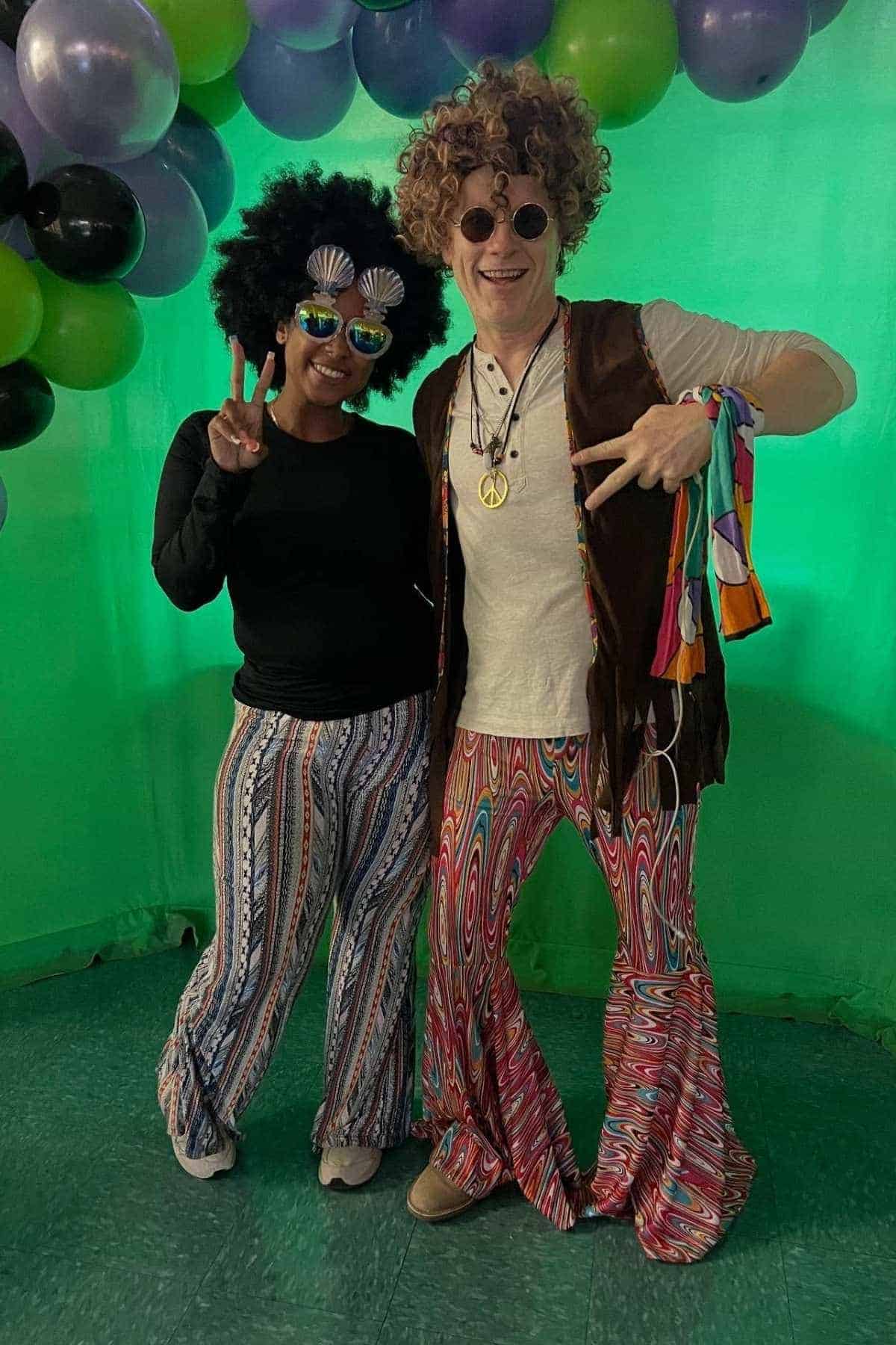 Two people dressed in 1970s-style clothing and wigs stand smiling under balloons, making peace signs.