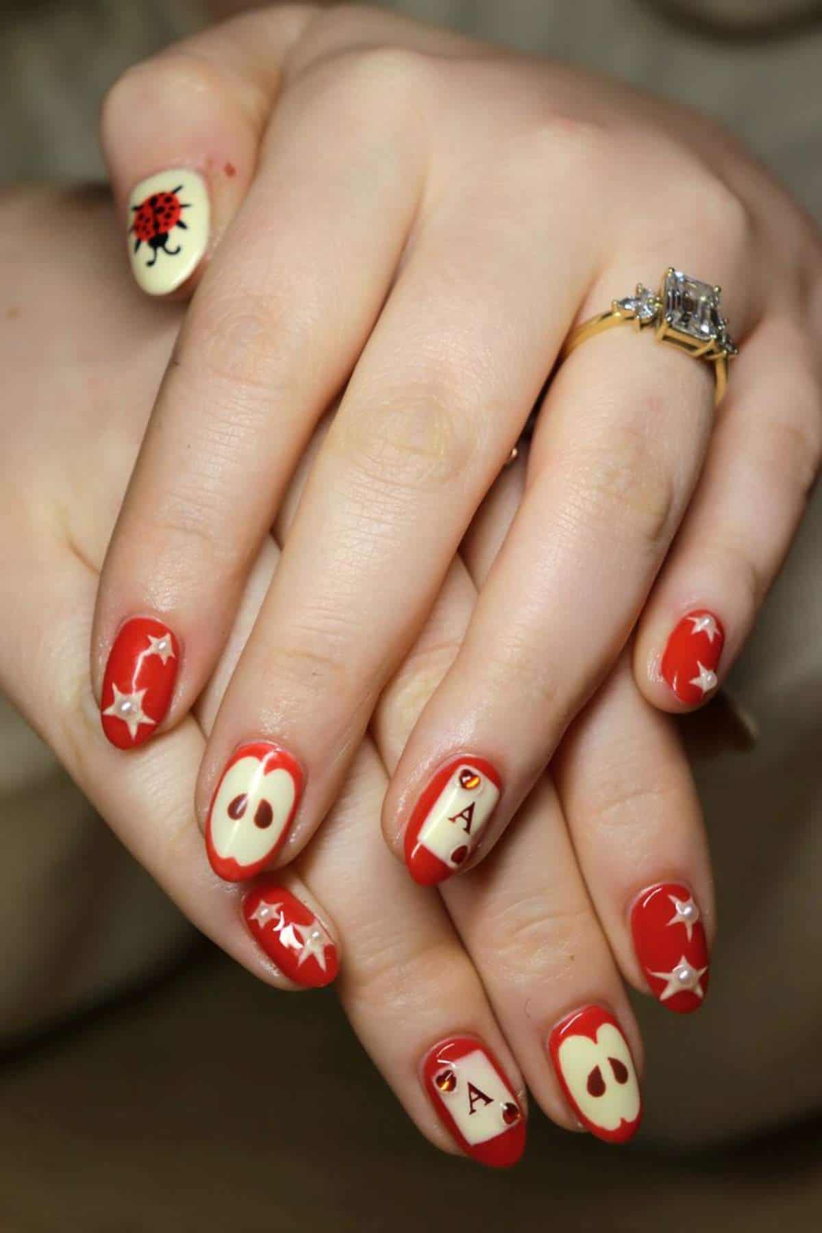 A person’s hands with red and cream nail art featuring playing card suits, aces, dice, and star designs. The person wears a gold ring with a rectangular gemstone.