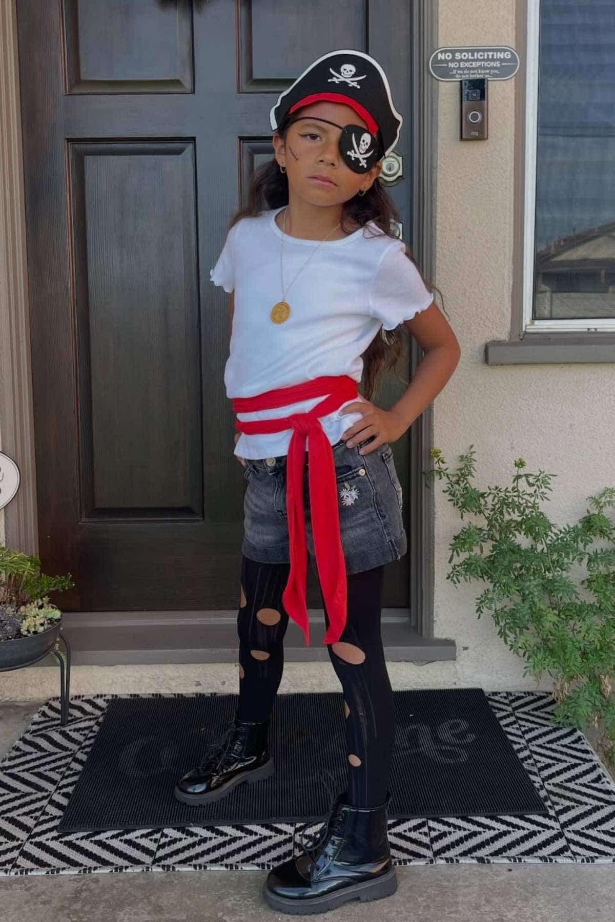 Child dressed as a pirate stands on a doorstep wearing a pirate hat, white shirt, red sash, black skirt with leggings, and black boots. A "No Soliciting" sign is visible on the door.