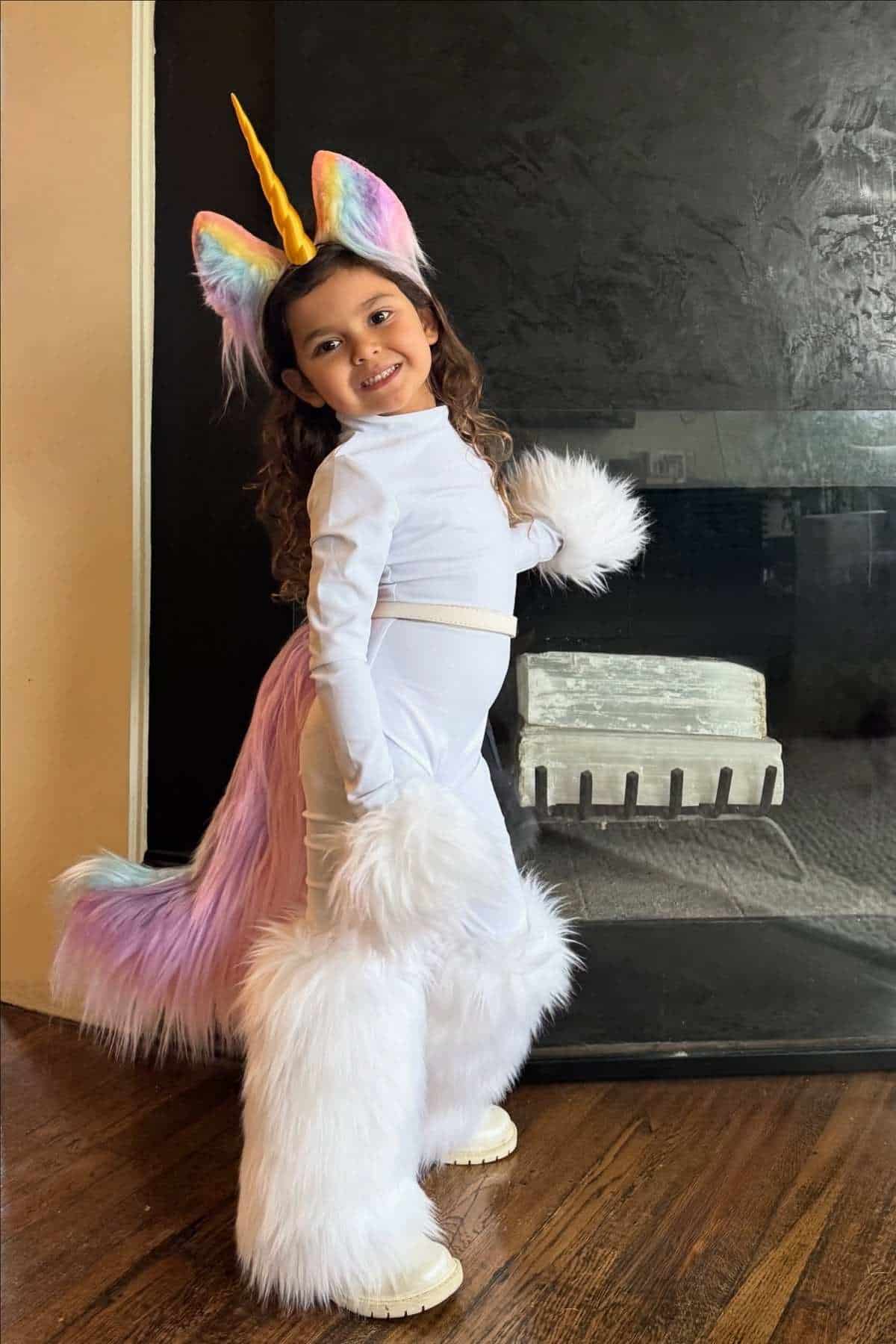 A young child in a unicorn costume with a yellow horn, rainbow ears, pastel tail, and furry white leg and arm warmers stands on a wooden floor, posing and smiling at the camera.