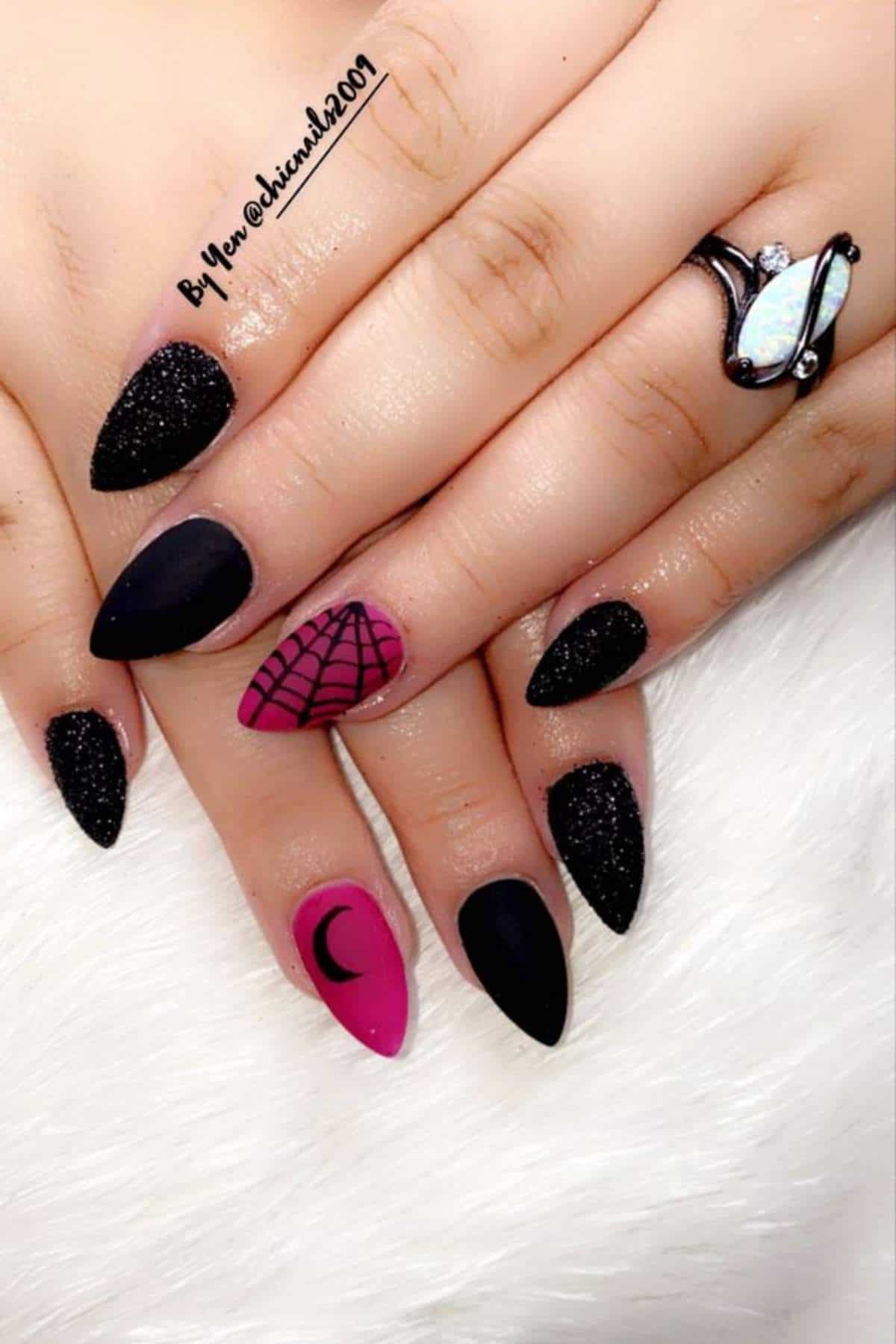 Hands with long, pointed nails in black and pink polish; designs include a crescent moon, net pattern, and glitter. A ring with an oval stone is worn on one finger.