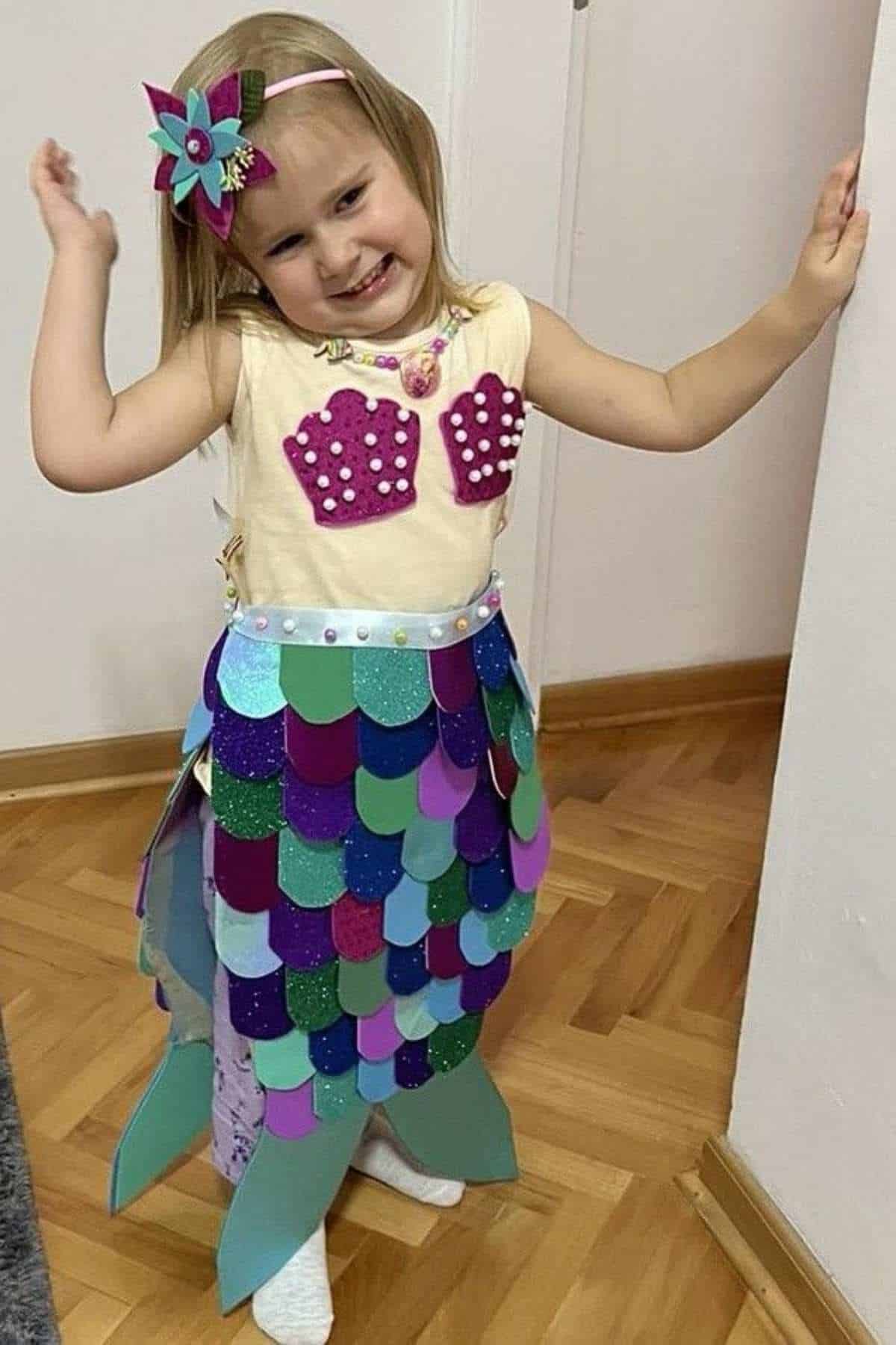 A young girl smiles and poses indoors, wearing a homemade mermaid costume with a seashell top, colorful tail, and a matching headband.