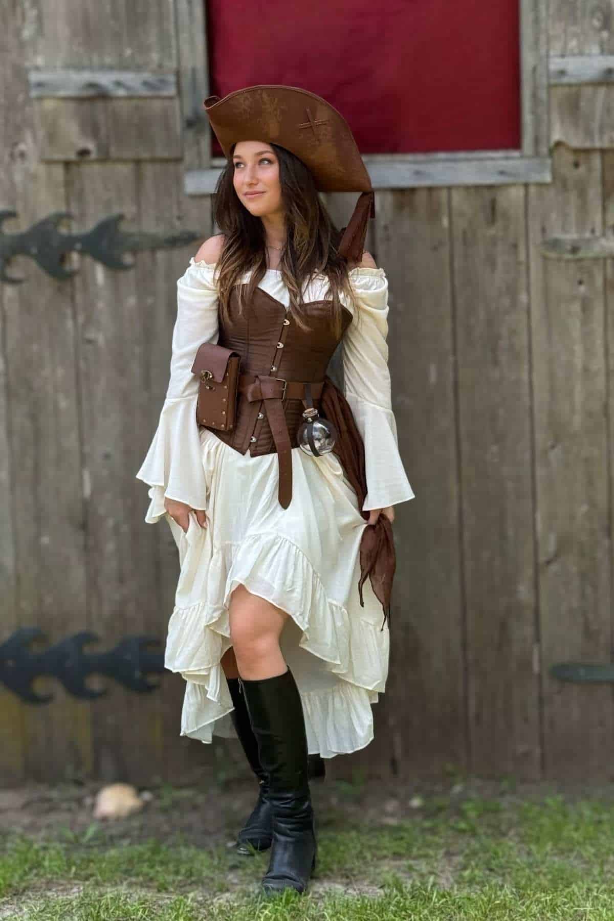 Woman in a brown and white pirate costume with boots and a hat stands outdoors in front of a wooden building.