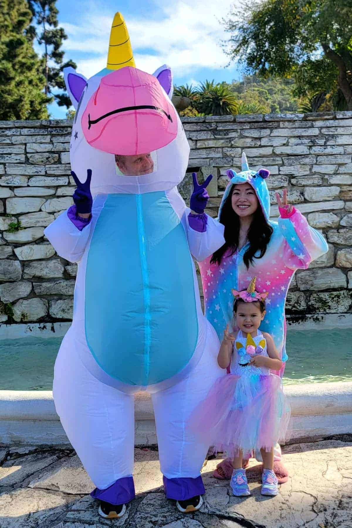 Three people dressed in unicorn-themed costumes pose and make peace signs in front of a stone wall and fountain outdoors.