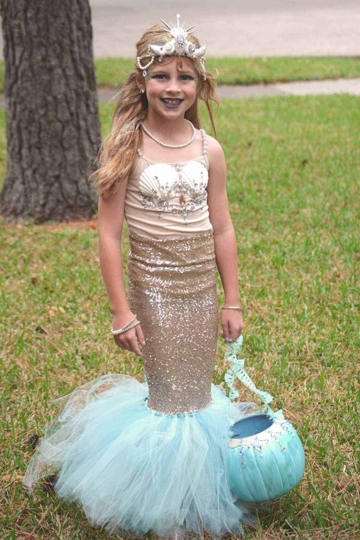 Young girl in a sparkly mermaid costume stands on grass, holding a blue pumpkin-shaped bucket. She wears a seashell headpiece and has long blond hair.