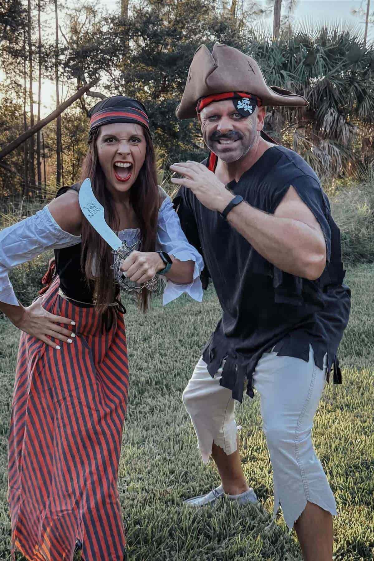 Two adults dressed as pirates pose outdoors; the woman holds a curved toy sword and the man wears a pirate hat with an eye patch and mustache.