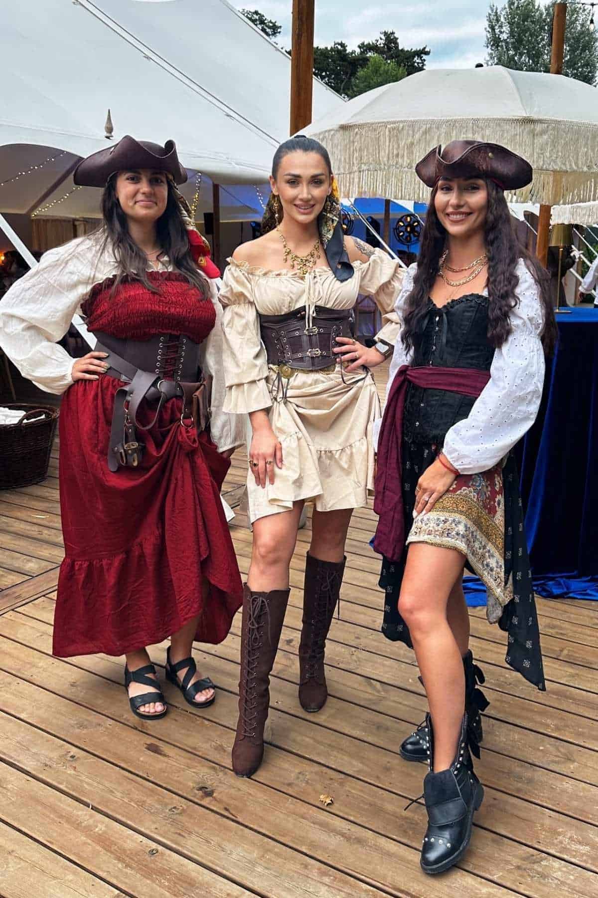 Three women wearing pirate-themed costumes pose together on a wooden deck at an outdoor event with tents in the background.