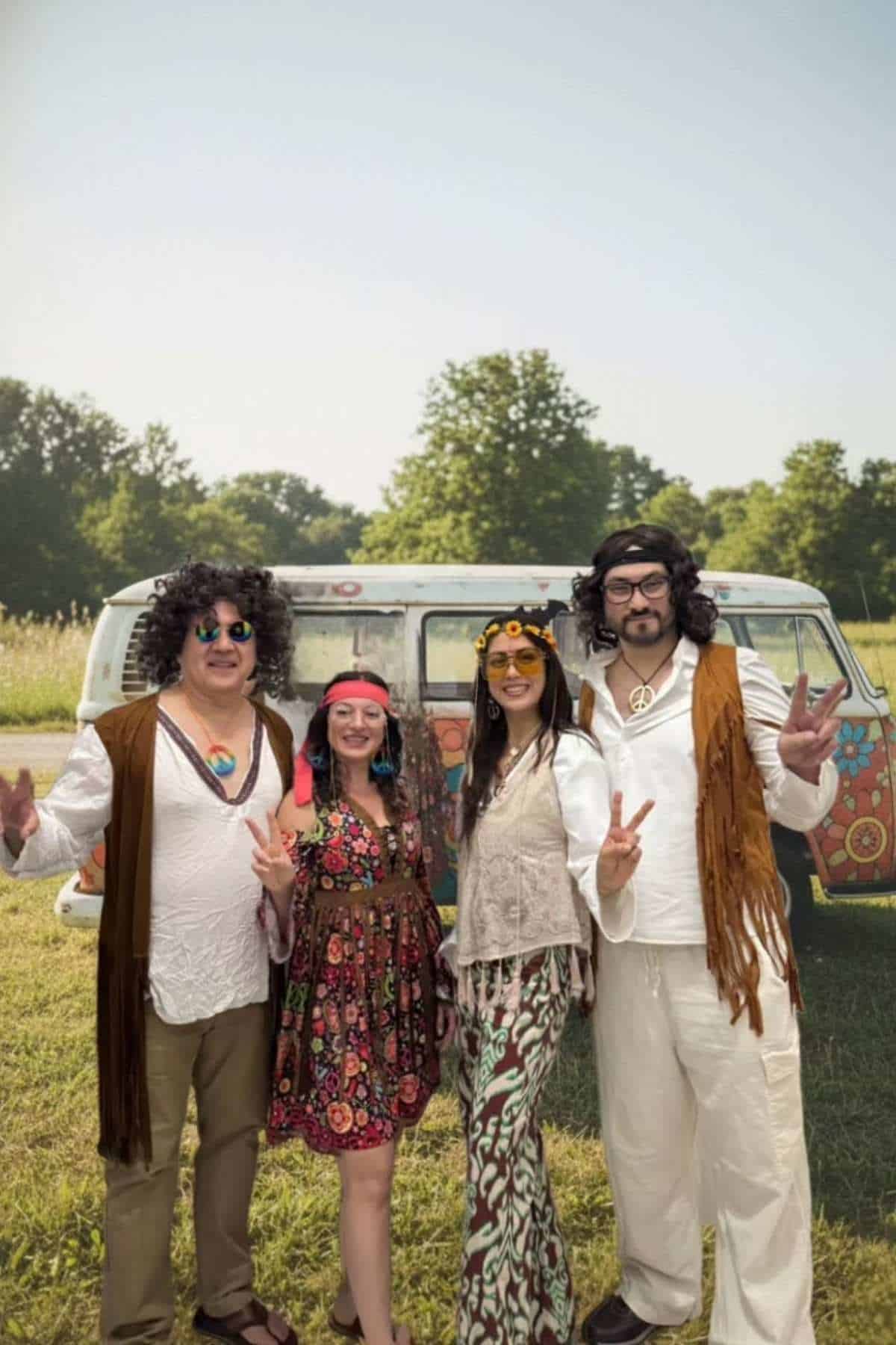 Four adults in retro hippie costumes pose outdoors in front of a decorated van, making peace signs with their hands.