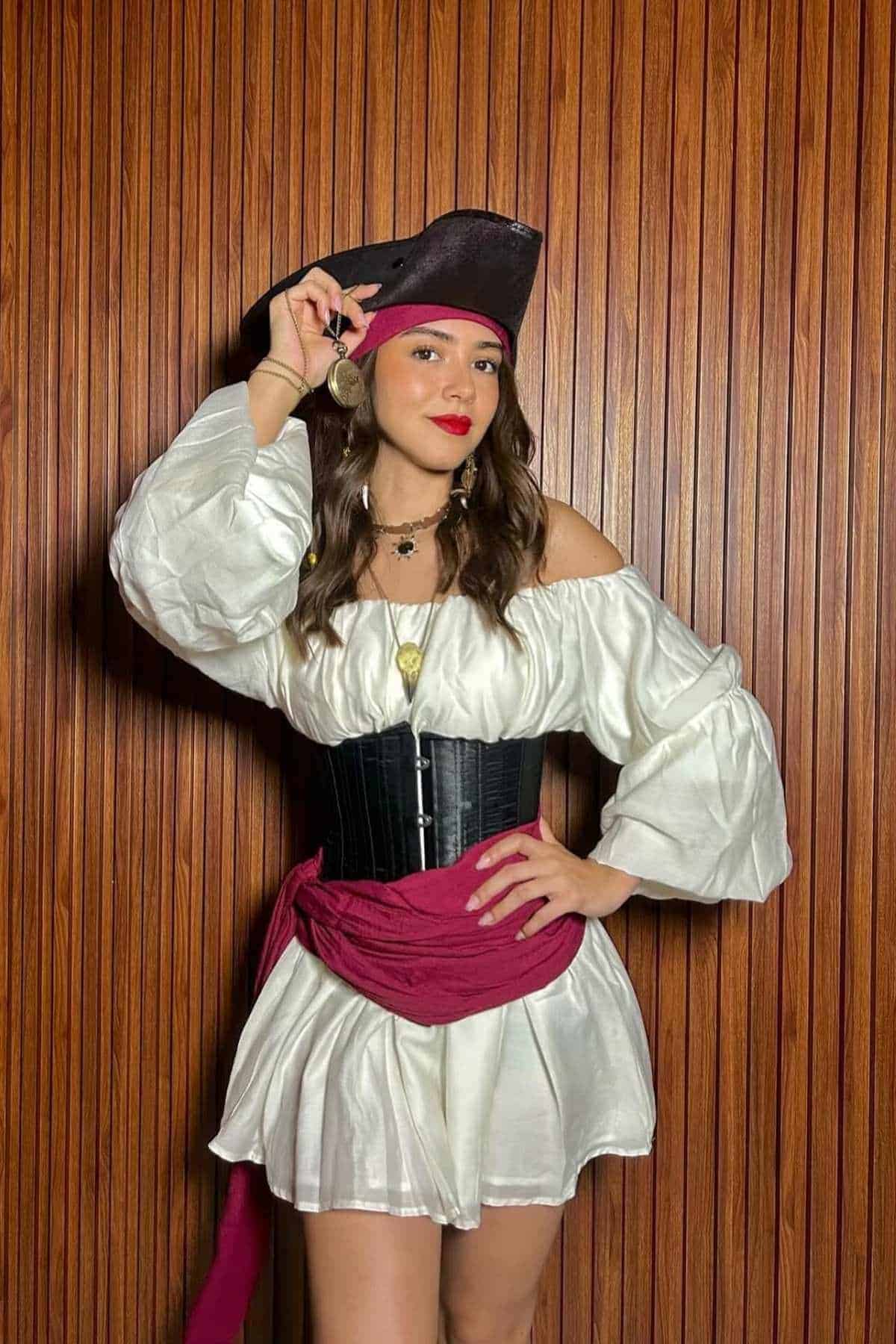 Woman in a pirate costume with a white off-shoulder dress, black corset, red sash, black hat, and gold earring, standing in front of a wooden wall.