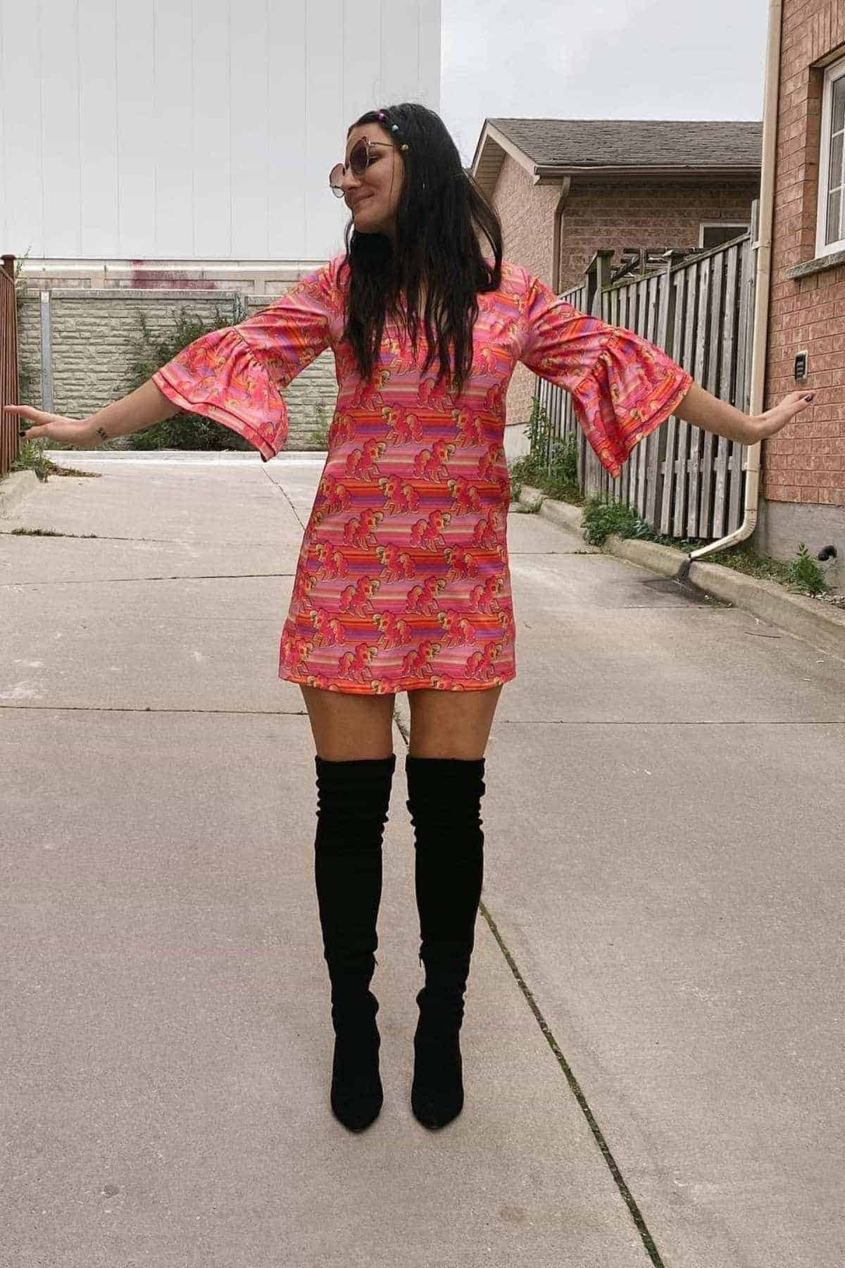 Woman stands outdoors with arms outstretched, wearing a pink printed dress with flared sleeves and black over-the-knee boots, sunglasses, and smiling.