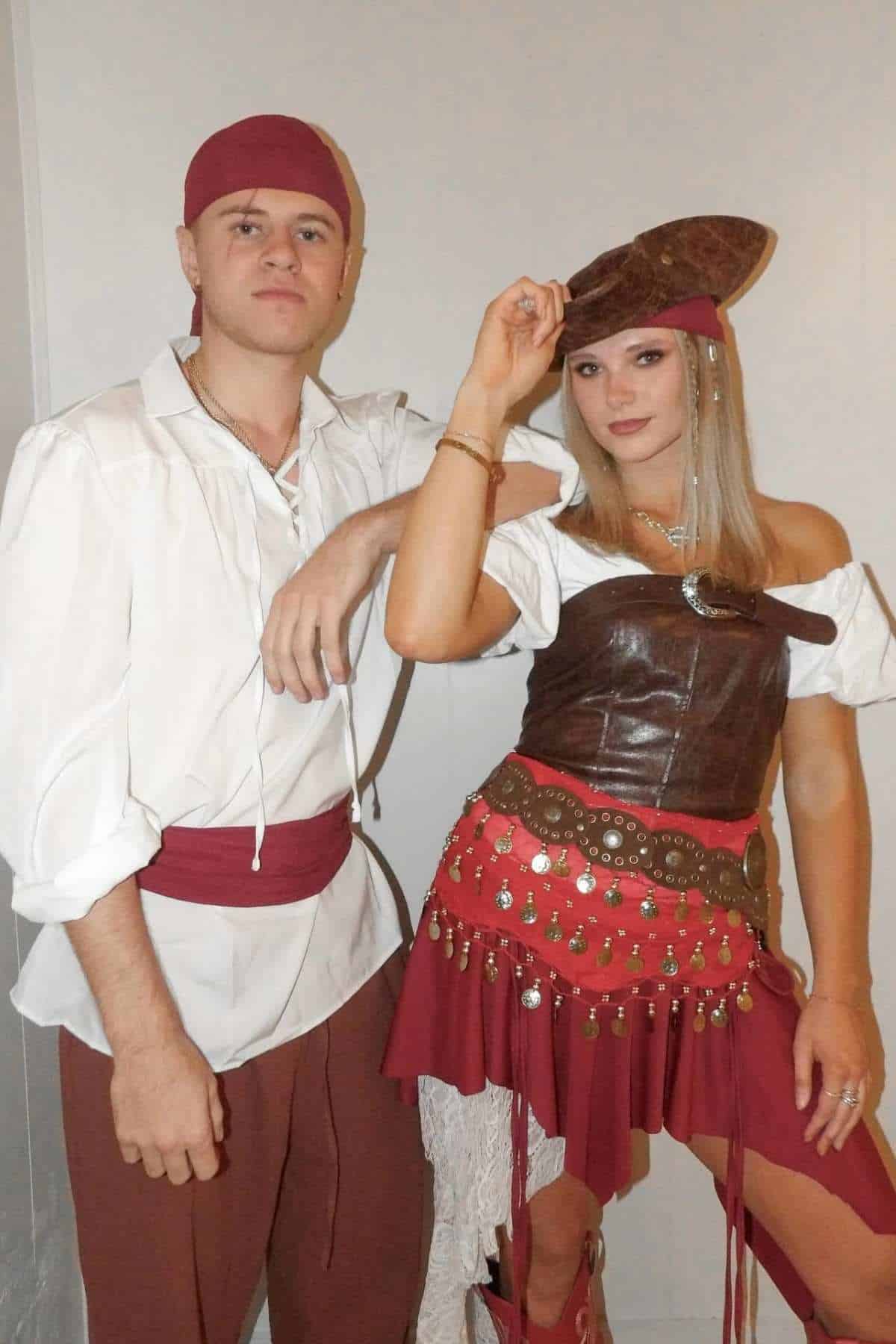 Two adults dressed in pirate costumes stand against a plain wall; the man wears a white shirt and red sash, and the woman wears a brown corset, red skirt, and brown pirate hat.