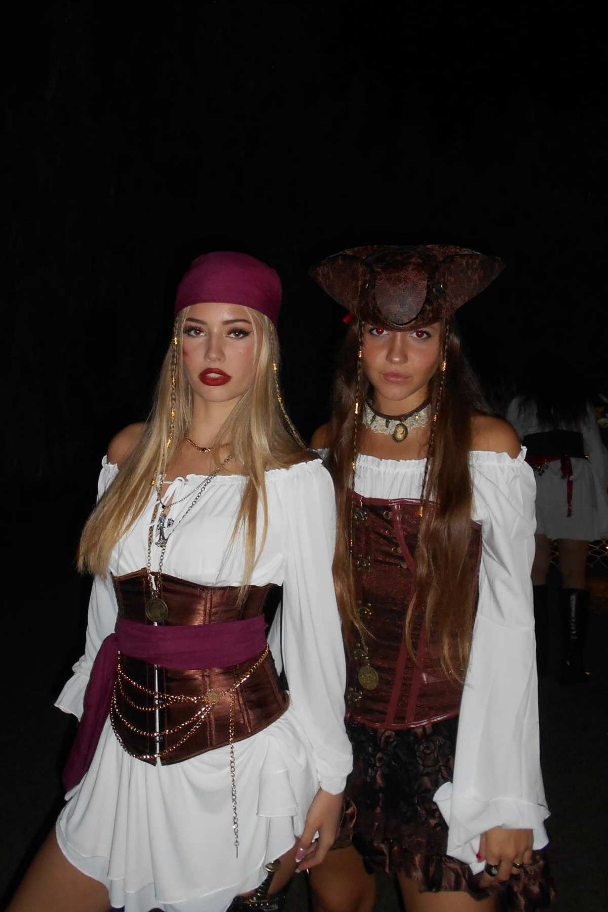 Two women dressed as pirates in white blouses, brown corsets, and accessories stand side by side against a dark background.