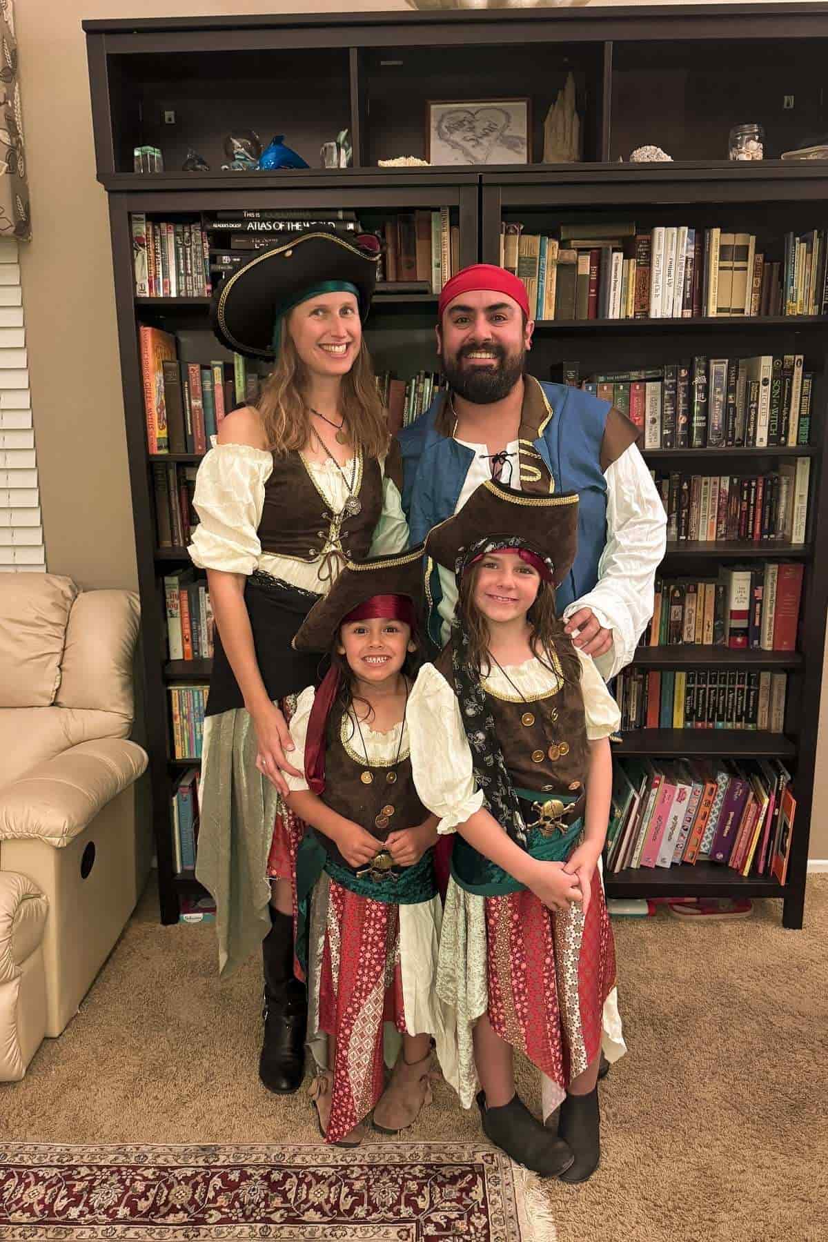 A family of four poses indoors dressed in coordinated pirate costumes, standing in front of a bookshelf.