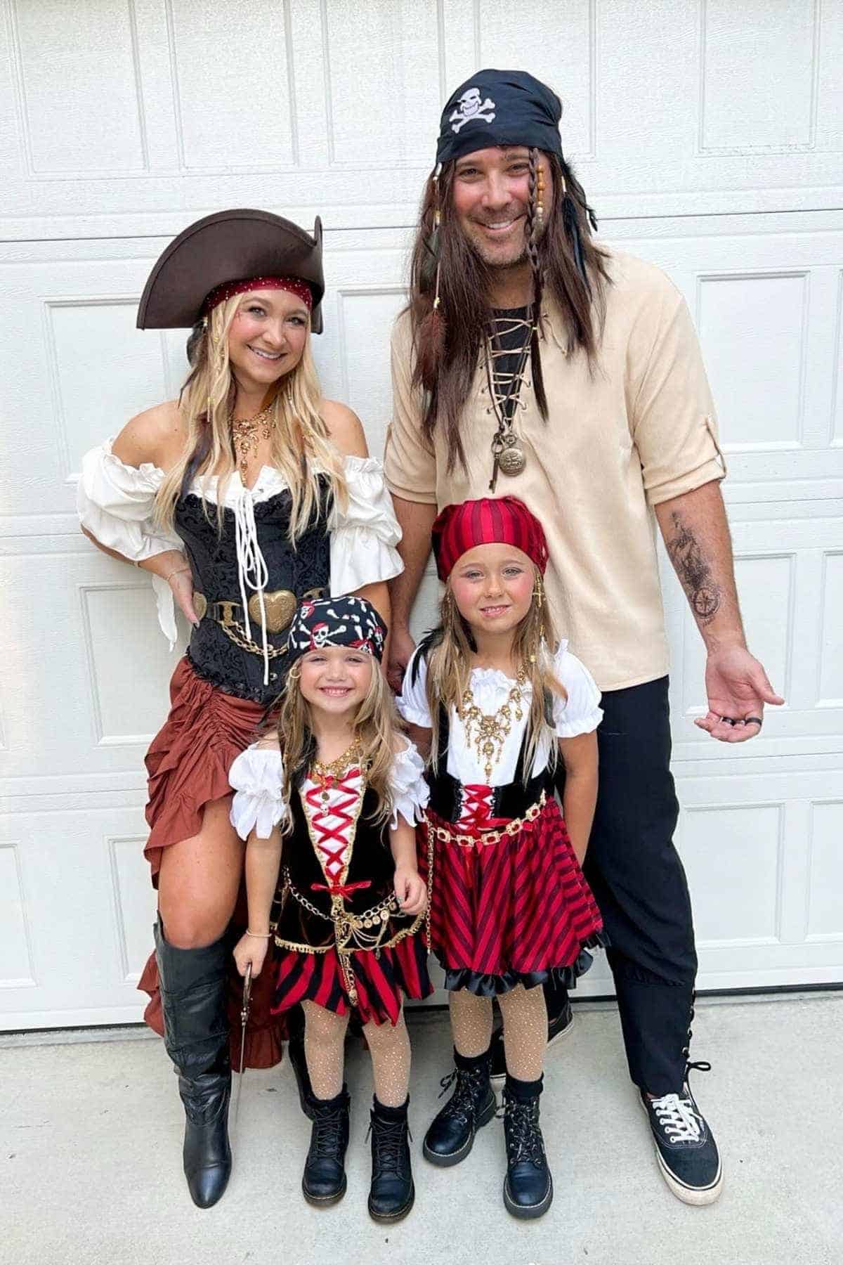 A family of four dressed in pirate costumes stands and smiles in front of a white garage door.