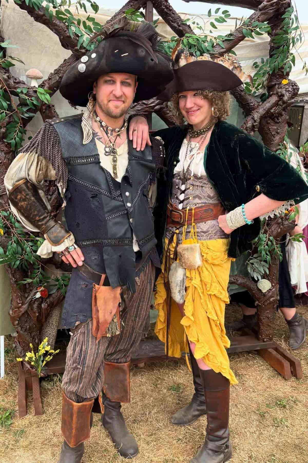 Two people in elaborate pirate costumes pose outdoors, standing on grass in front of a leafy decorative backdrop at what appears to be a festival or fair.