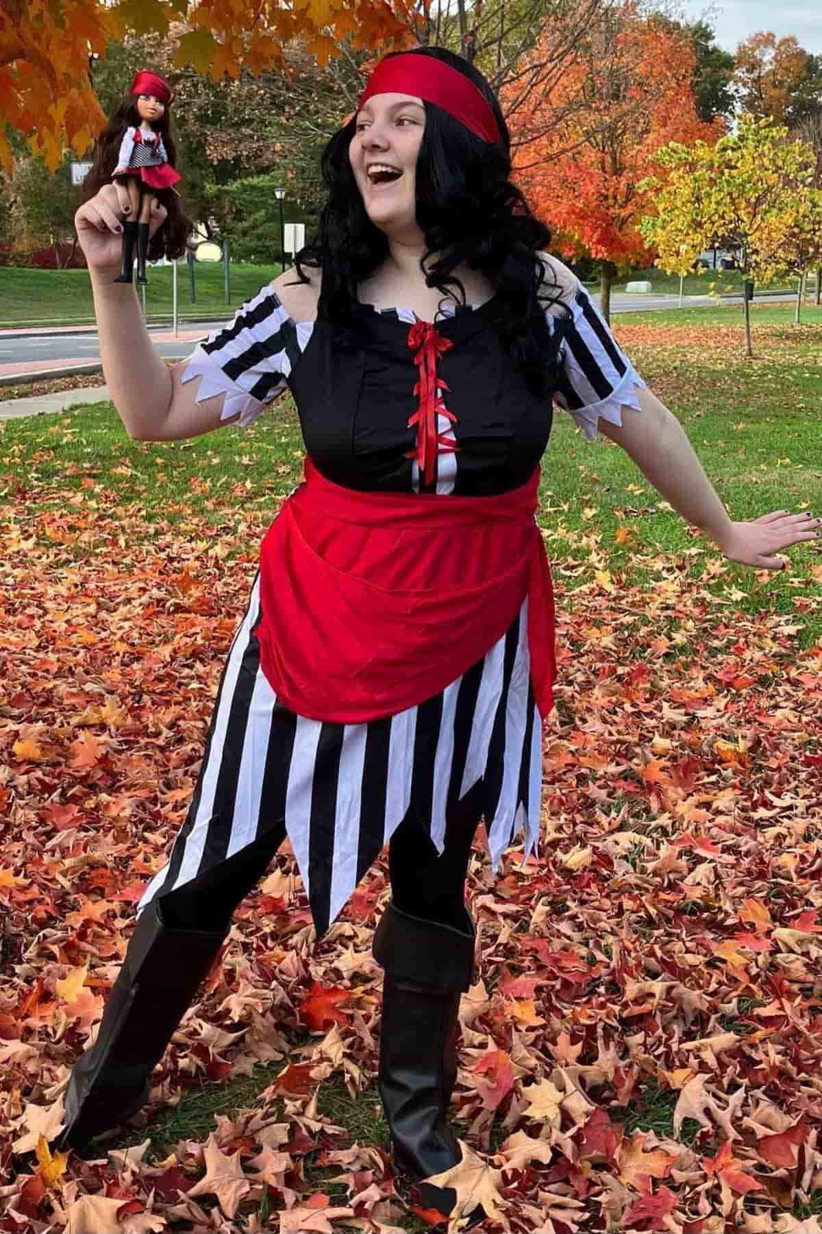 Person in a pirate costume stands on fallen autumn leaves, smiling and holding a small pirate doll aloft. Colorful trees and a street are visible in the background.