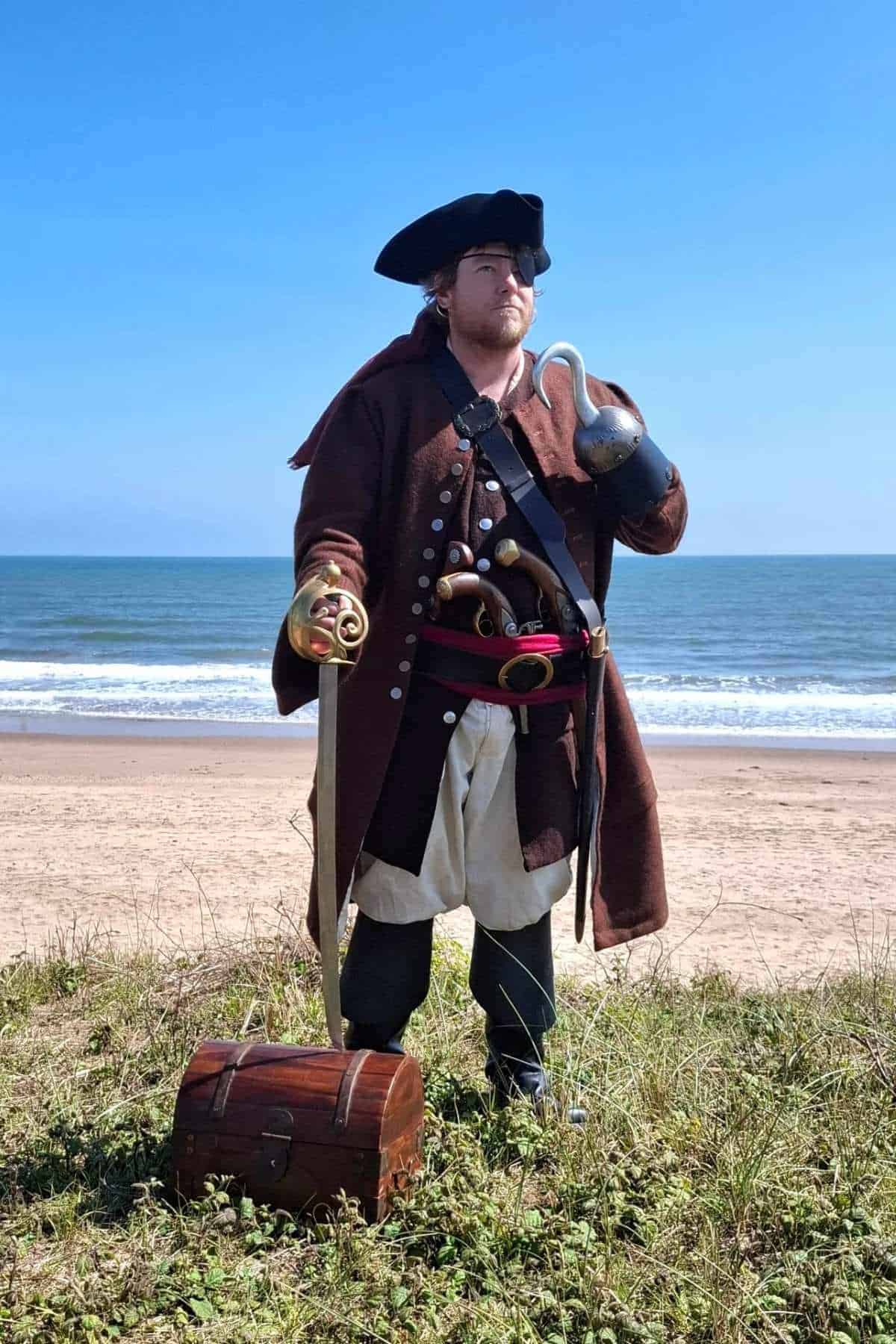 A person dressed as a pirate stands by the ocean, holding a sword and a hook, with a treasure chest on the grass in front.