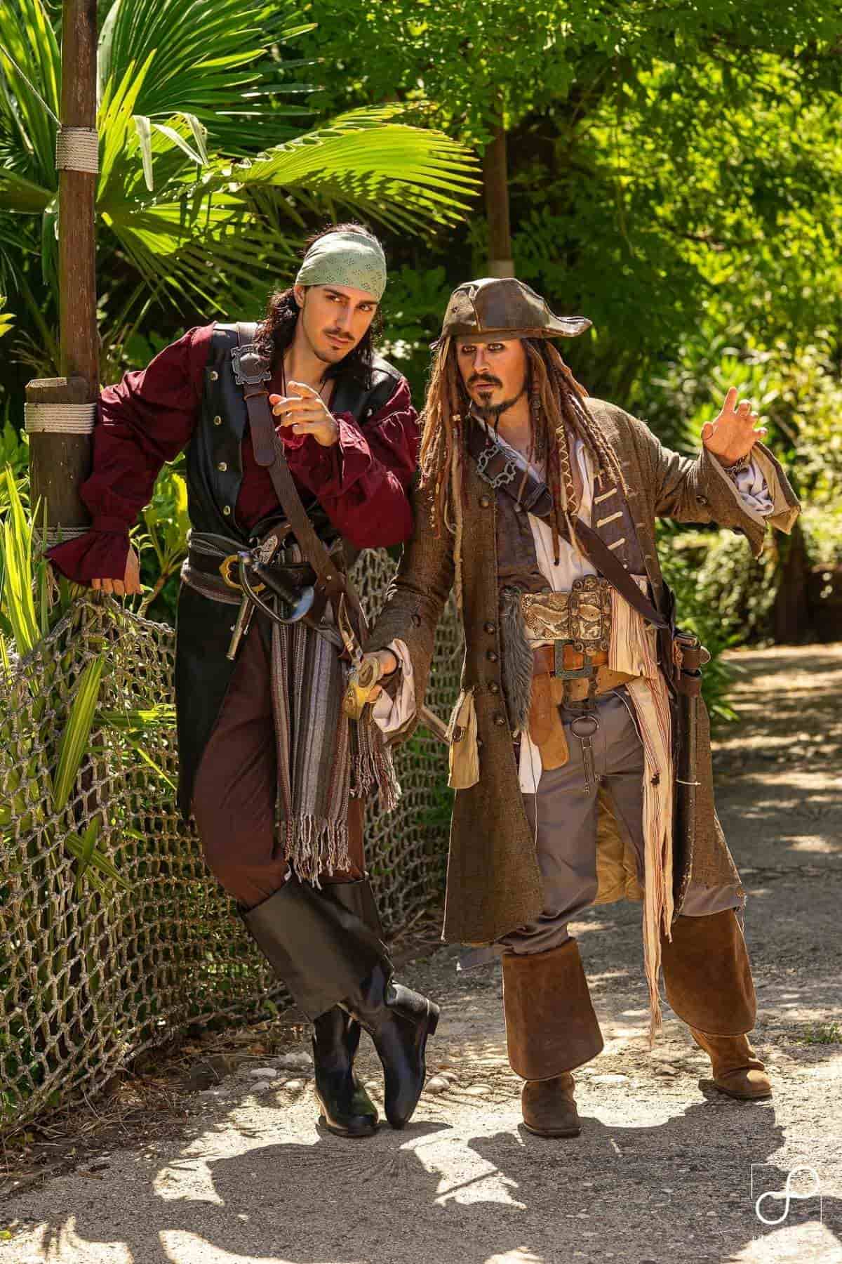 Two people dressed as pirates stand outdoors on a sunny day, leaning against a rope fence with greenery in the background.