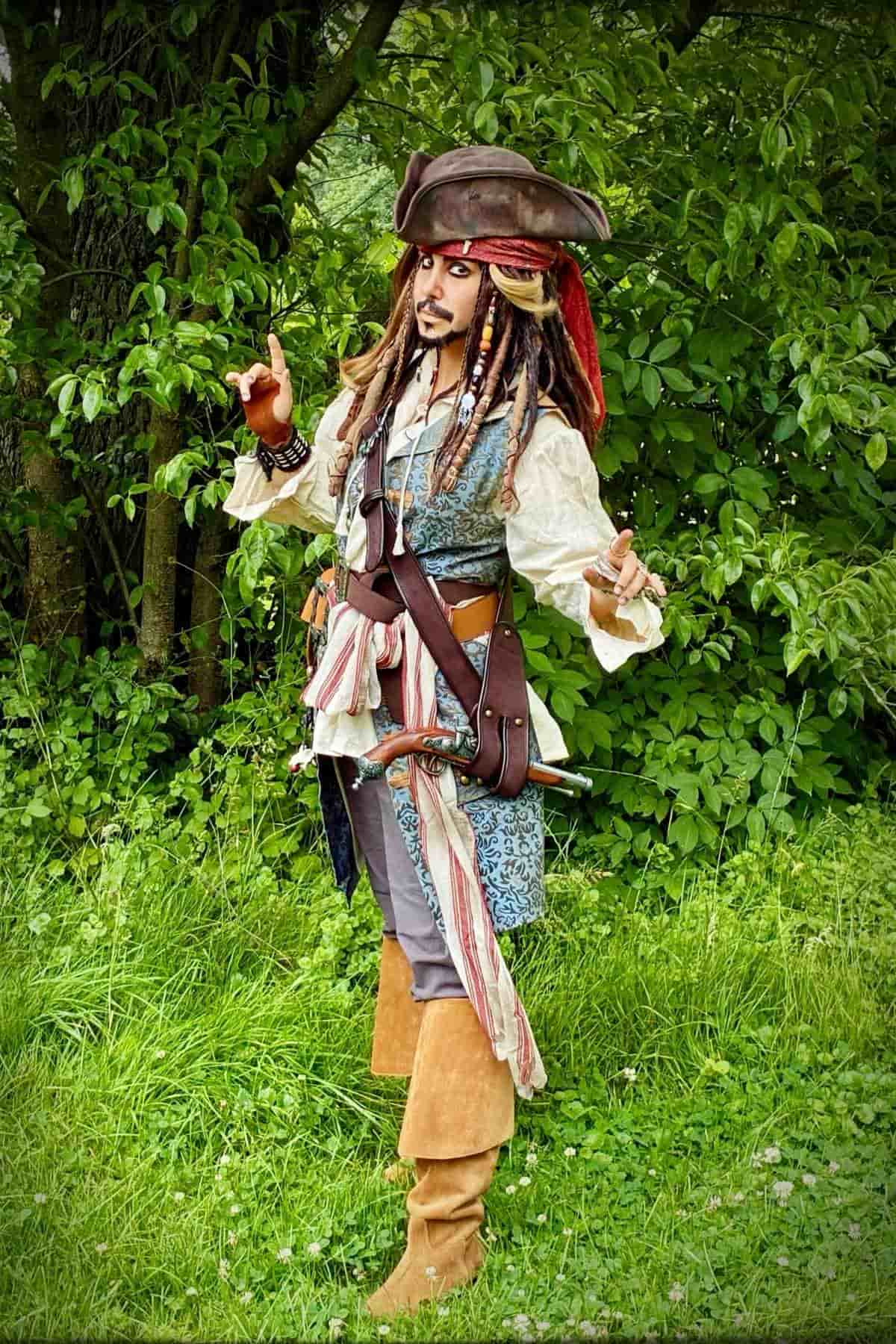Person in a pirate costume stands outdoors on grass, holding an object in one hand, surrounded by green foliage.