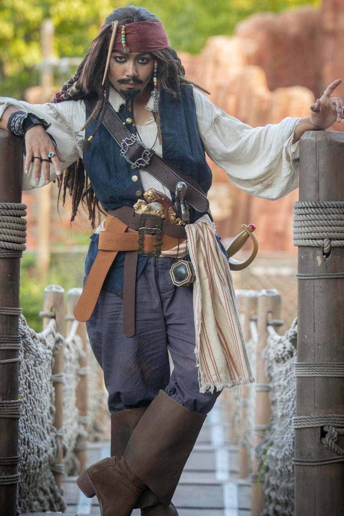 Person dressed as a pirate stands on a wooden bridge, wearing a tricorn hat, vest, boots, and accessories, with one hand resting on the rail and the other pointing.