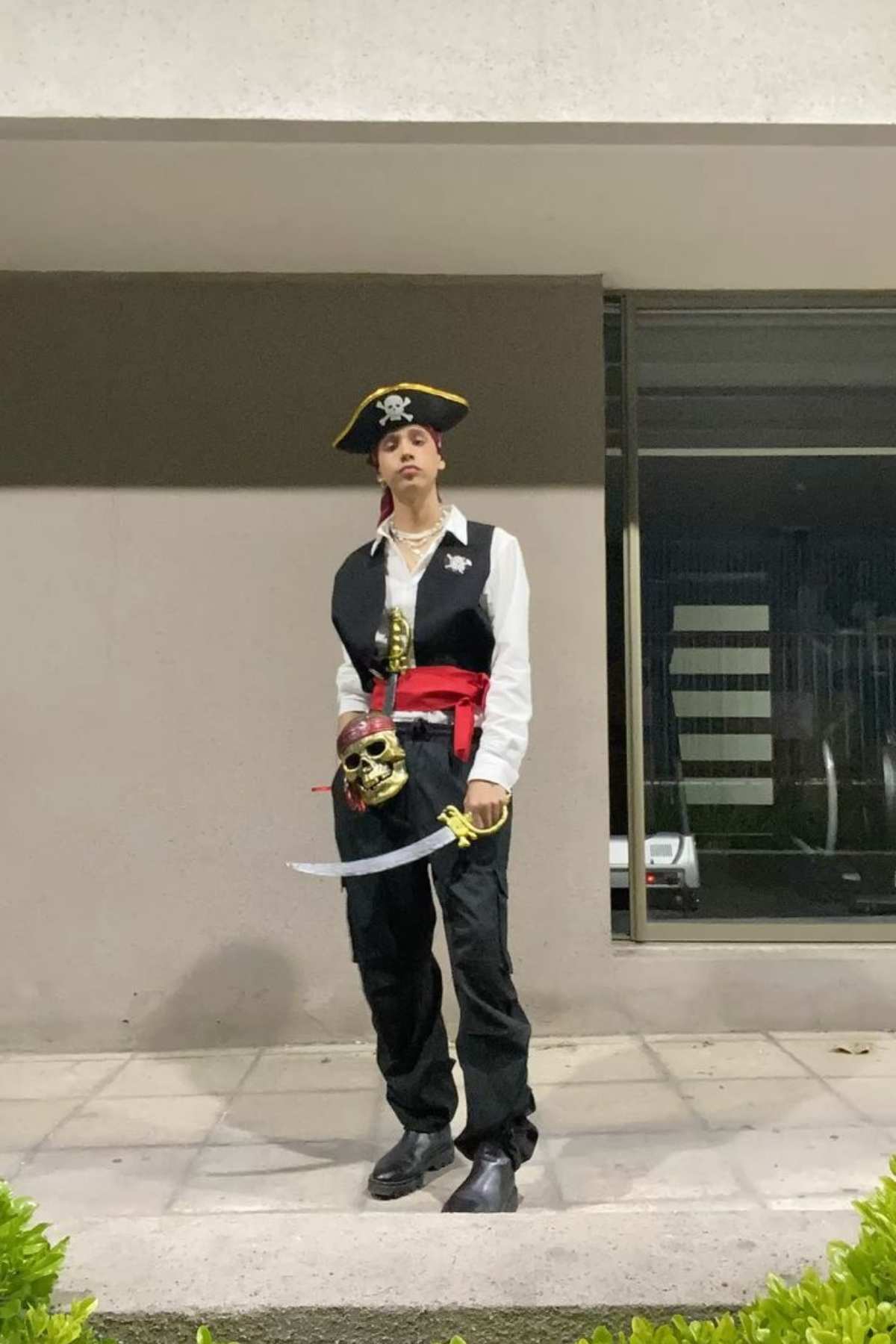 Person dressed as a pirate, wearing a black hat with a skull, white shirt, black vest, red sash, and holding a toy sword, standing on a sidewalk in front of a building.