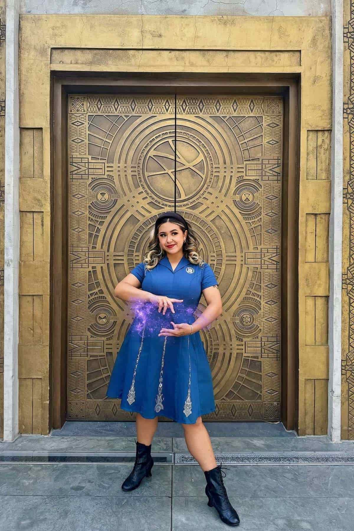Person in a blue dress and black boots stands in front of a decorative golden door, holding their hands as if conjuring purple glowing energy.