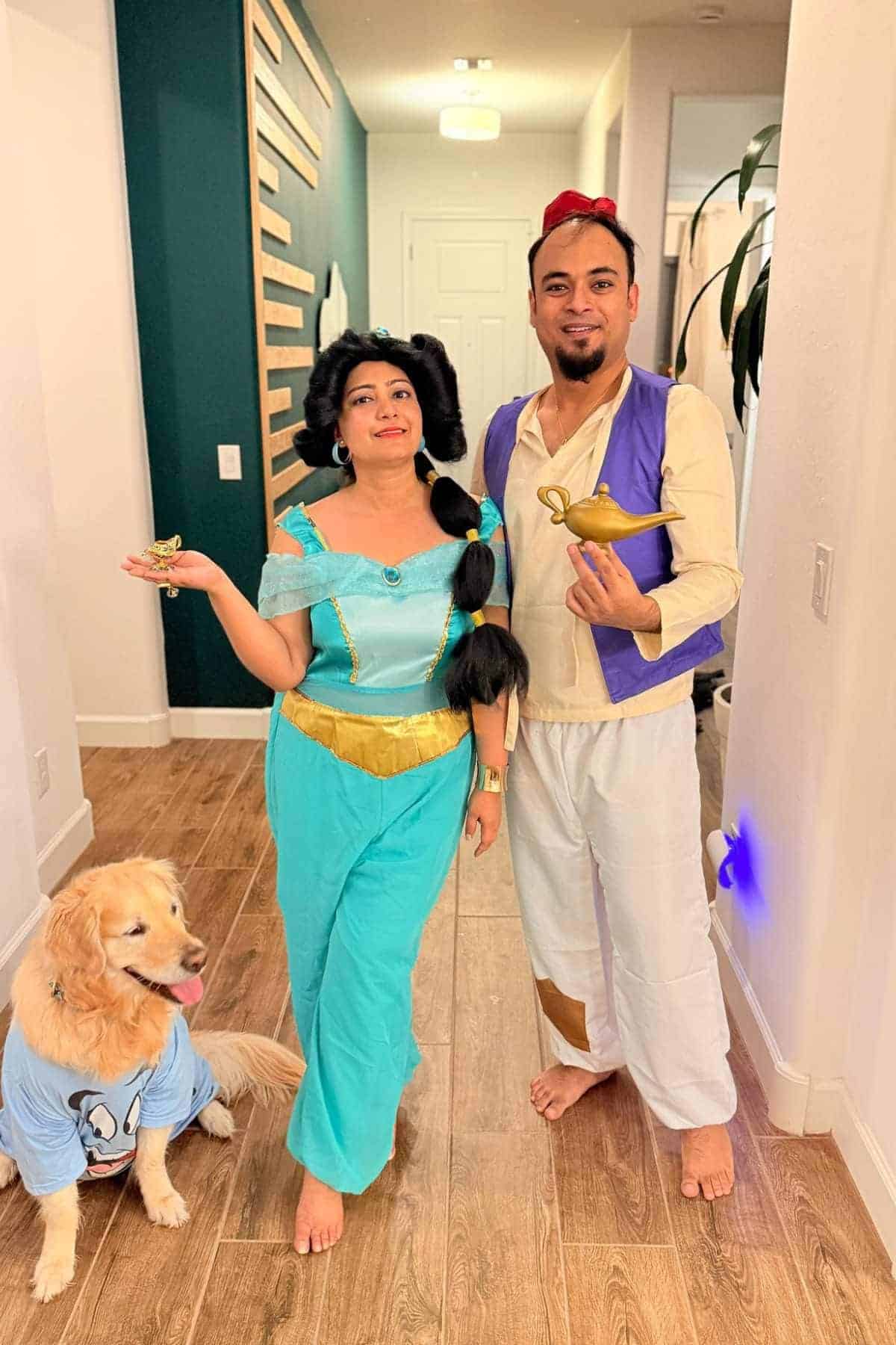 Two adults dressed as Jasmine and Aladdin hold props, while a golden retriever in a blue shirt sits beside them in a hallway with wood flooring.