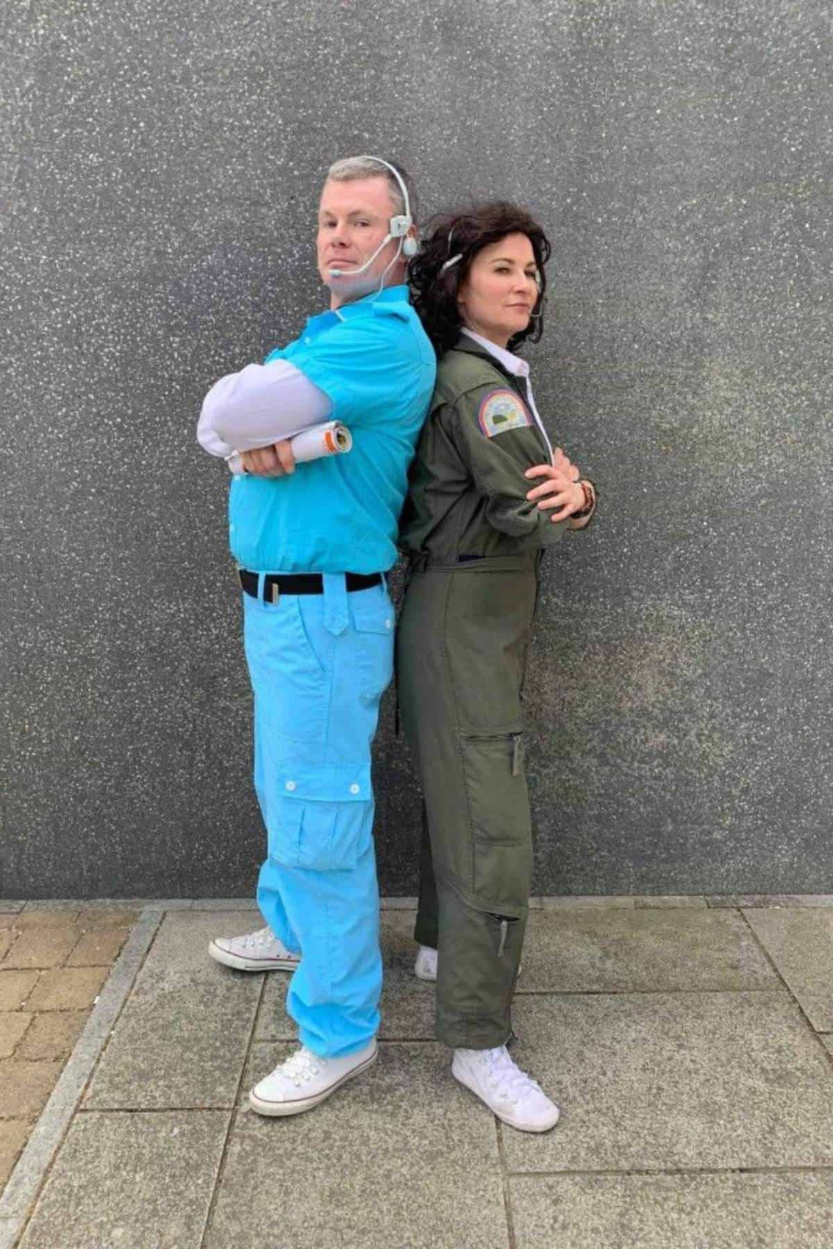 Two people stand back-to-back; one wears a light blue outfit and headset, the other wears a green jumpsuit with patches. Both have arms crossed and face opposite directions.