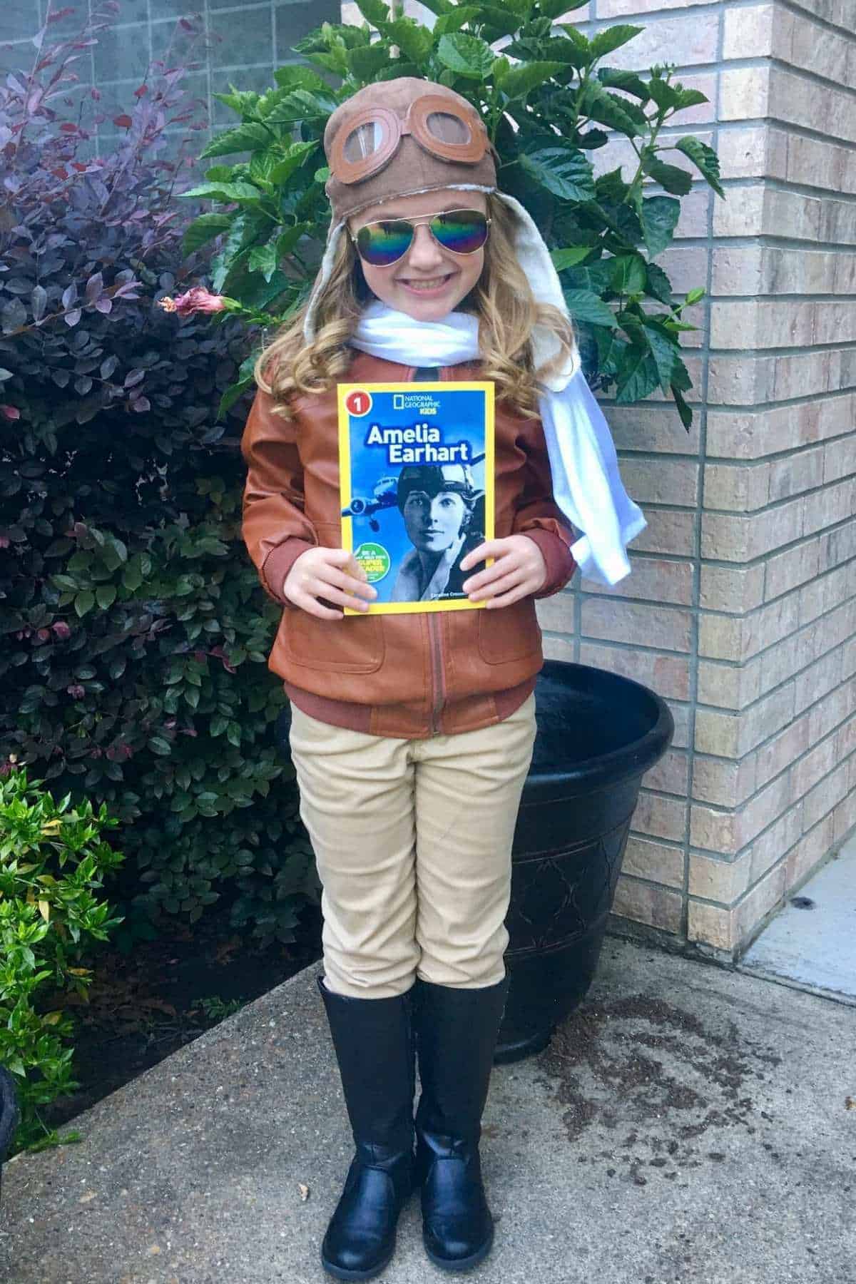 A child in an aviator costume stands outside, smiling and holding a book titled "Amelia Earhart.