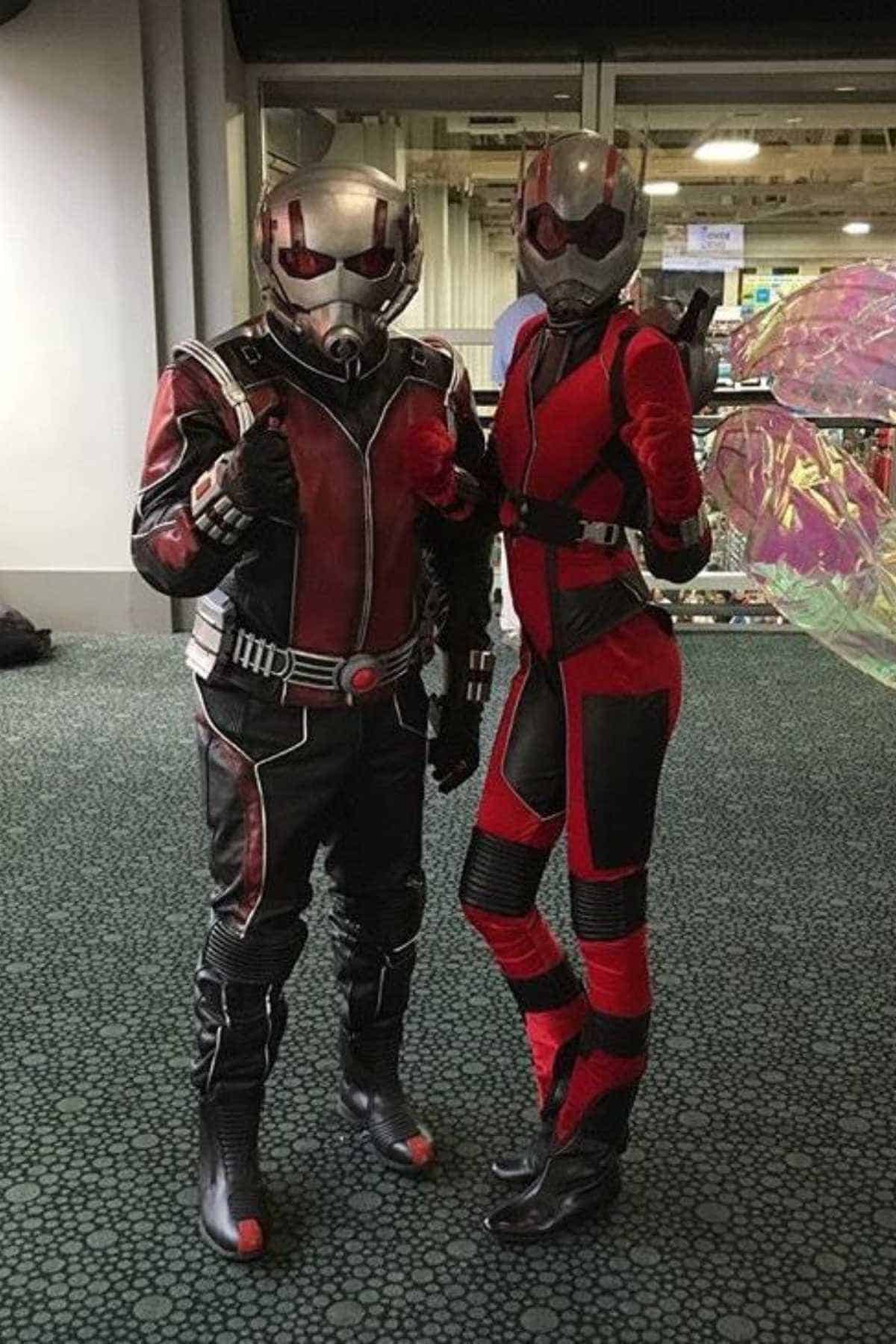 Two people dressed in Ant-Man and Wasp costumes pose together indoors, standing on a carpeted floor with glass windows and people in the background.