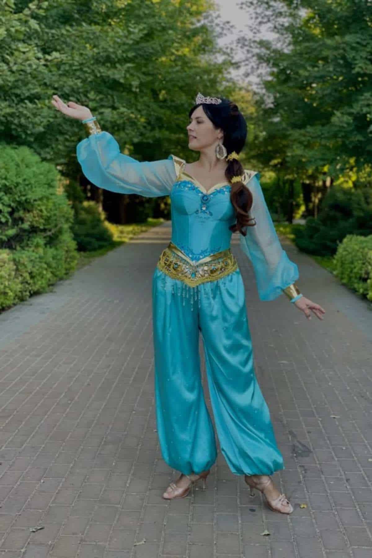 Person wearing a blue costume with gold accents and a tiara stands on a brick path in a park, raising one arm and looking to the side.