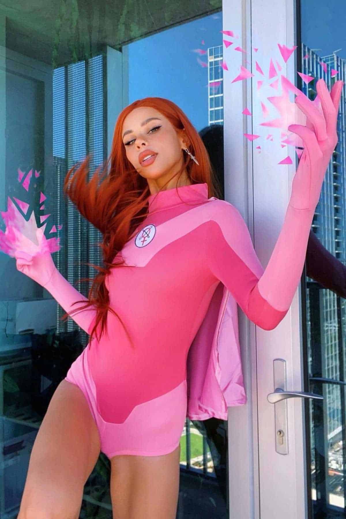Person in a pink superhero costume poses indoors with digital pink energy effects emanating from their hands near a glass door and city view.