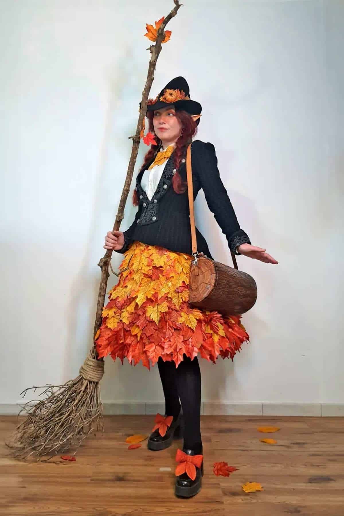 Person dressed in a costume with a skirt of autumn leaves, black jacket, hat, and holding a broomstick, standing indoors on a wooden floor.