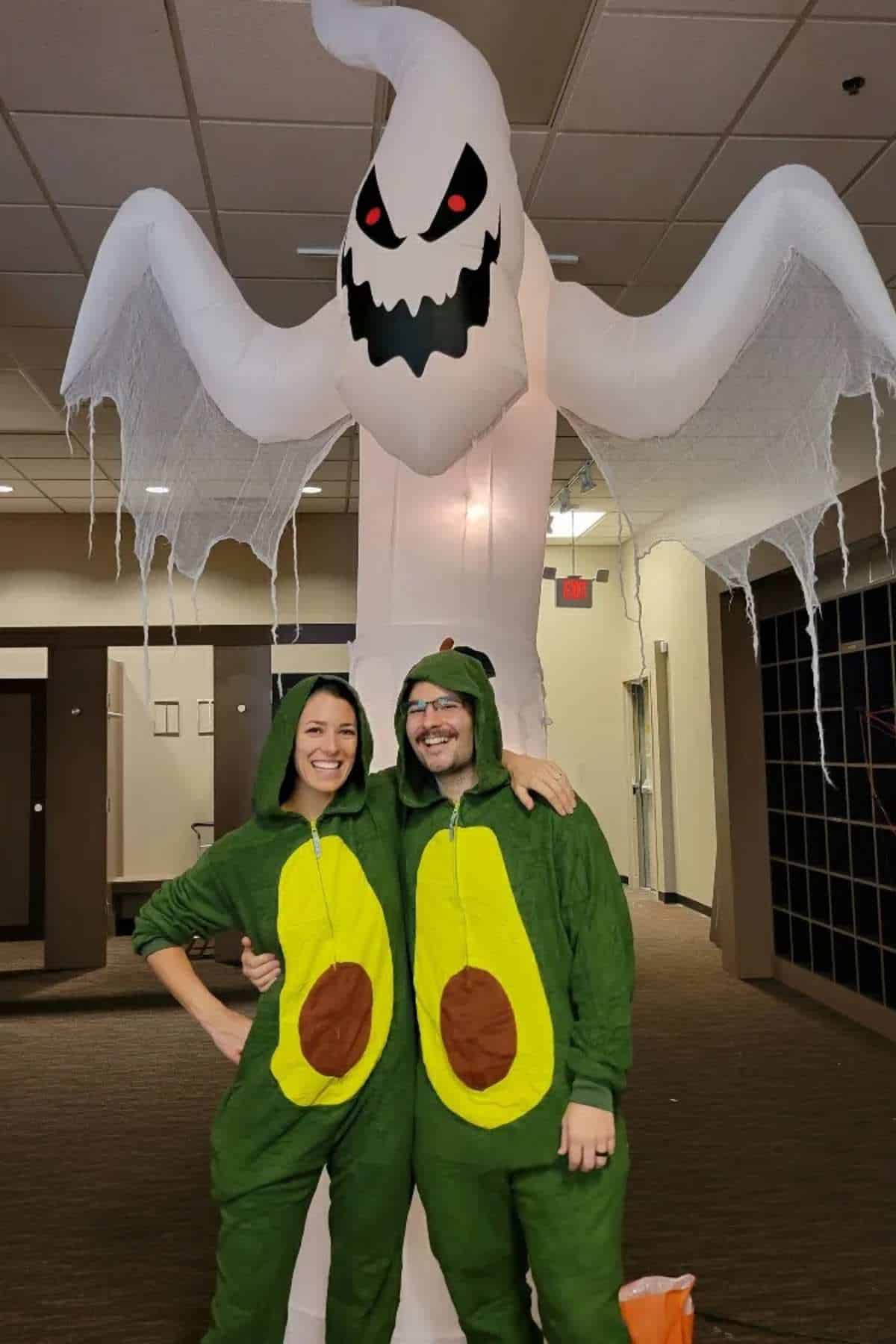 Two people in matching avocado costumes stand arm in arm in front of a large inflatable ghost decoration indoors.