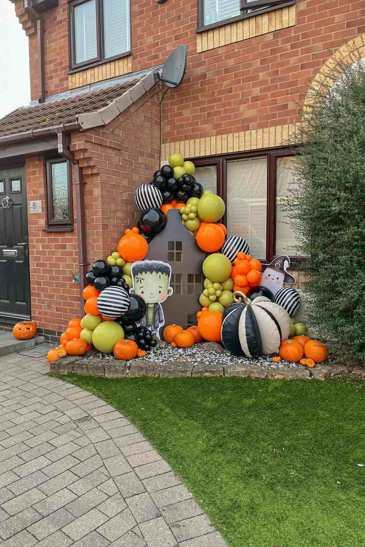 Halloween yard display with orange, green, and black balloons, pumpkins, a Frankenstein figure, and a haunted house backdrop in front of a brick home.