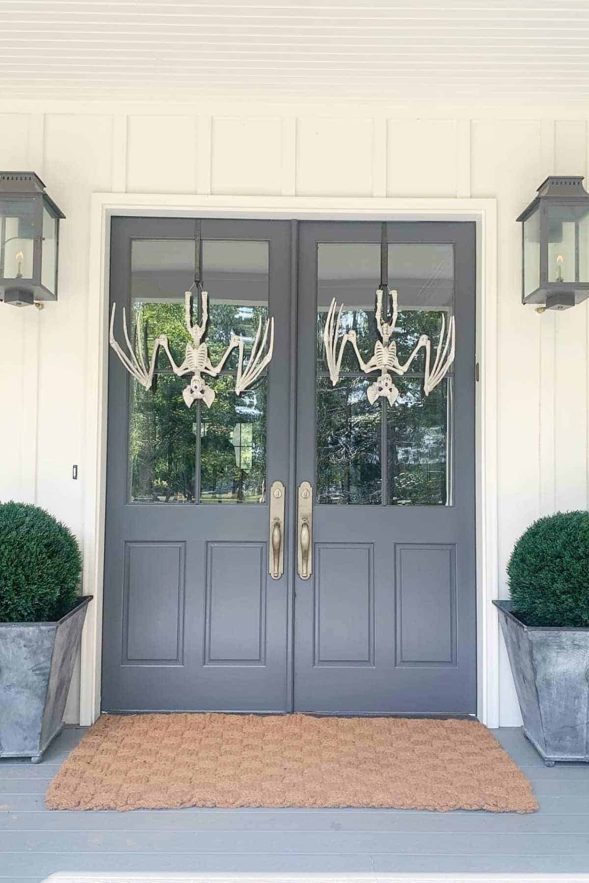 Double gray doors with glass panels have two bat-shaped Halloween decorations hanging, flanked by topiary shrubs in planters and lantern-style lights on each side, with a tan doormat in front.
